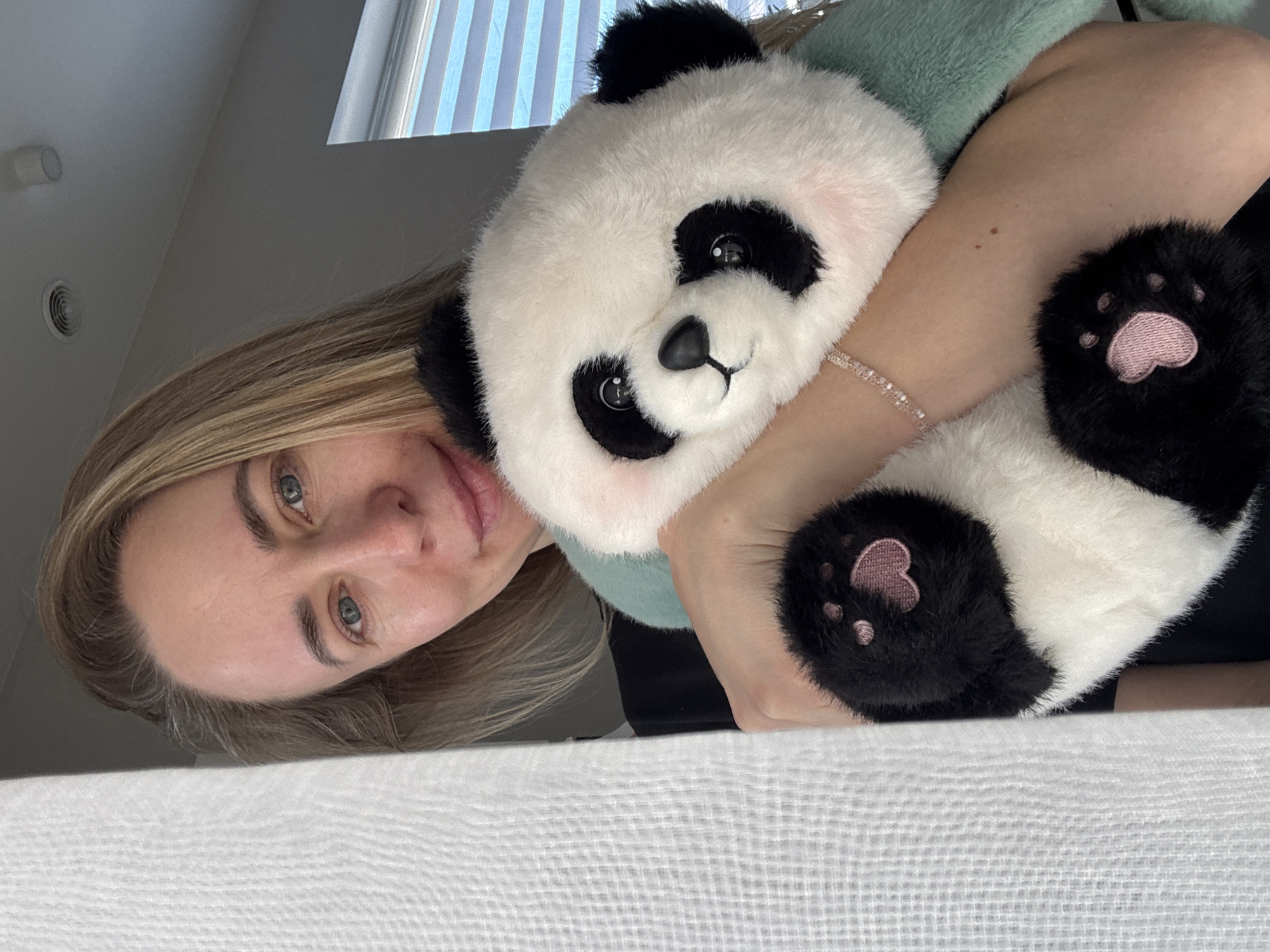 A selfie shows a blonde woman holding a panda plush toy. The woman has light skin and blue eyes. She is wearing a bracelet. The panda toy is white and black with pink paw pads. A pale green scarf is around the panda’s neck. The background includes a window with blinds and a white surface with a textured pattern, likely a blanket or a bed. The location is not evident in the image. The subject is indoors. There is no evidence to suggest the location is Chaikovsky, Russia.