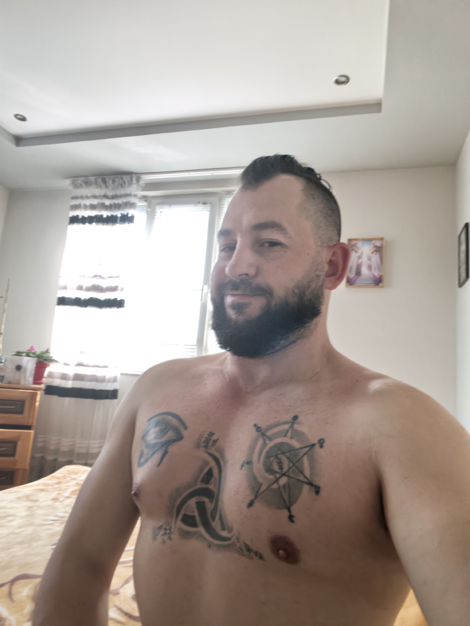 A shirtless man, appearing to be in his 30s or 40s, with short, dark hair and a full beard, is captured in a close-up, chest-up shot. He looks directly at the camera with a slight, friendly smile.
He has several tattoos on his chest. On his left pectoral (viewer's right), a large tattoo features an eye symbol, possibly the Eye of Horus, intertwined with intricate knotwork. On his right pectoral (viewer's left), there is a circular tattoo containing a seven-pointed star (heptagram) with smaller symbolic elements or markings around its perimeter, all within a larger shaded circle.
The man is indoors, likely in a bedroom or living room. Bright natural light streams in from a large window on the left side of the frame, partially covered by striped curtains in white, grey, and black. Below the window, a wooden dresser or nightstand holds a small red potted plant and a white container. The foreground shows part of a patterned blanket or bedspread in shades of light brown and gold.
On the wall to the man's right, a framed picture hangs, depicting what appears to be an ethereal or religious scene with glowing figures and archways. The ceiling is white with recessed lighting.
The image appears to be a daytime selfie, given the bright illumination and direct gaze of the subject. No discernible text is visible in the image, other than the stylized markings within the tattoos. The overall mood is calm and casual.