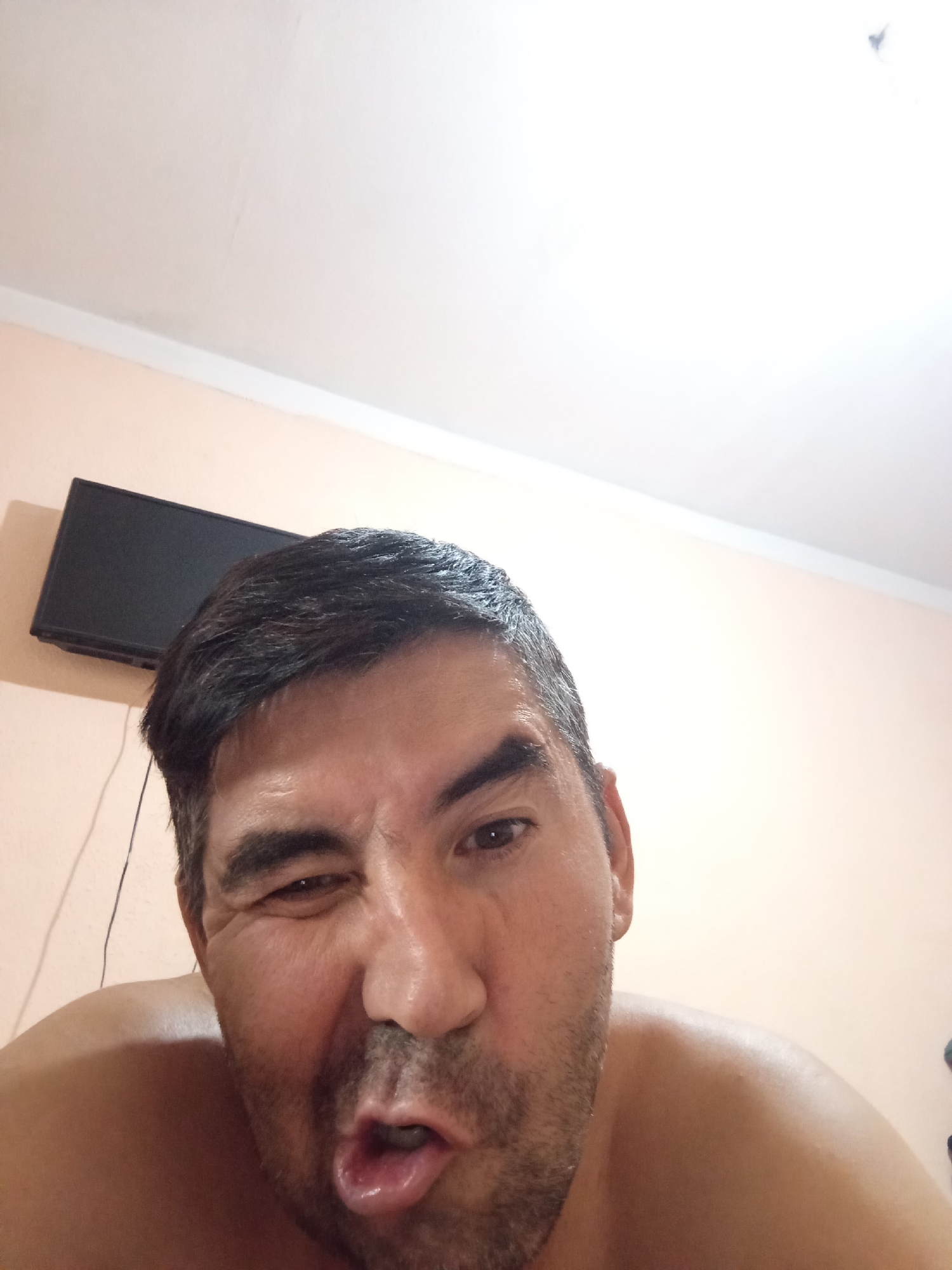 A man is shown in a selfie, winking with his right eye and sticking out his tongue slightly. He has dark hair and stubble. A television is mounted on a wall behind him. The room appears to be a bedroom, with a white ceiling and light peach walls. The image is taken from a low angle, looking up at the man.