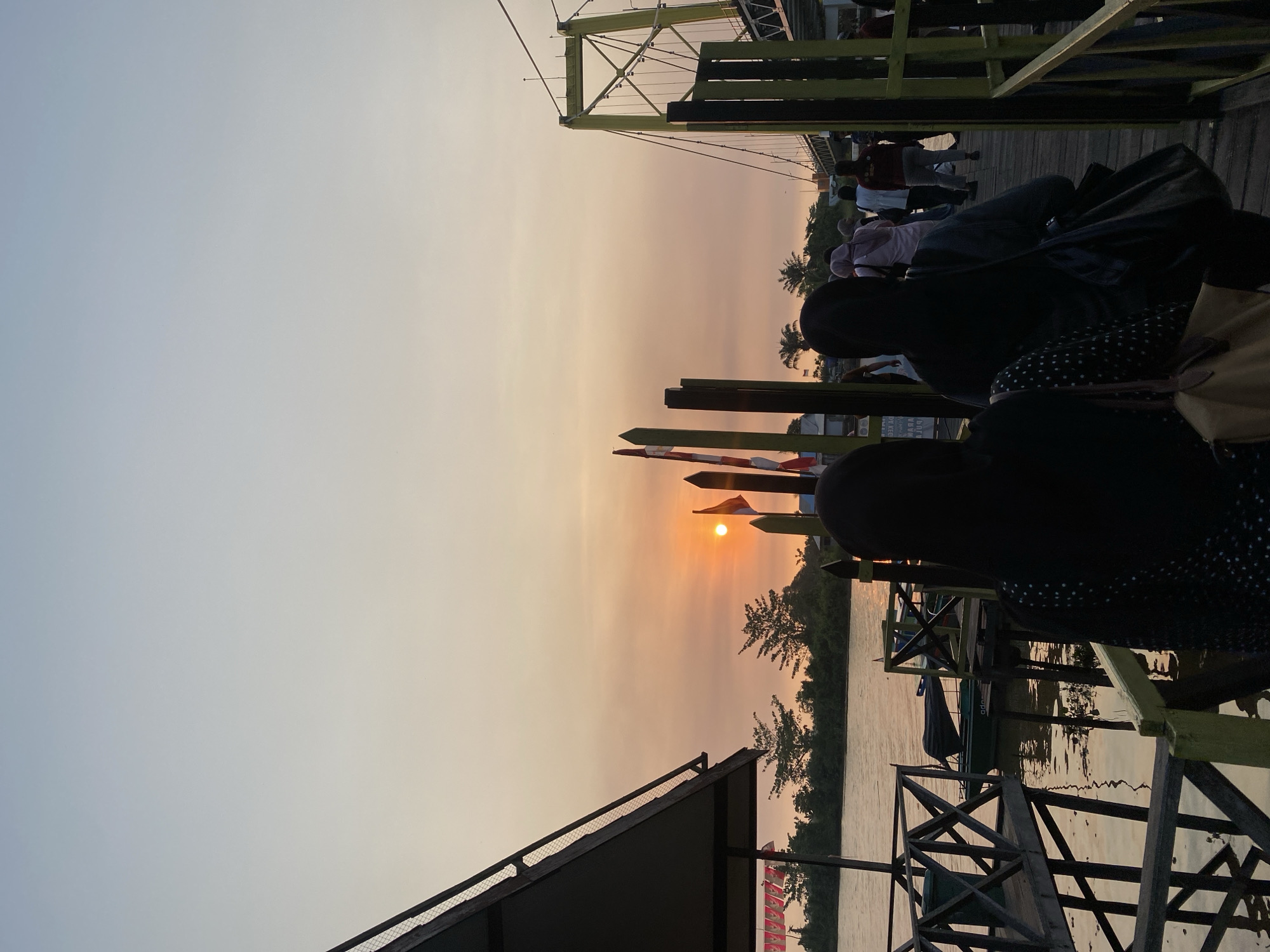 At sunset in Sungai Pitung, Indonesia, a group of people walks along a wooden pier towards a yellow suspension bridge. The sun, a bright orange orb, is visible low in the sky, casting a warm glow over the scene and reflecting on the water. Flags are planted along the pier, some of which appear to be national flags. Trees and vegetation line the opposite bank of the river. Several boats are moored along the pier. The perspective is from behind the walking figures, emphasizing the forward movement and the grand vista of the sunset and bridge.