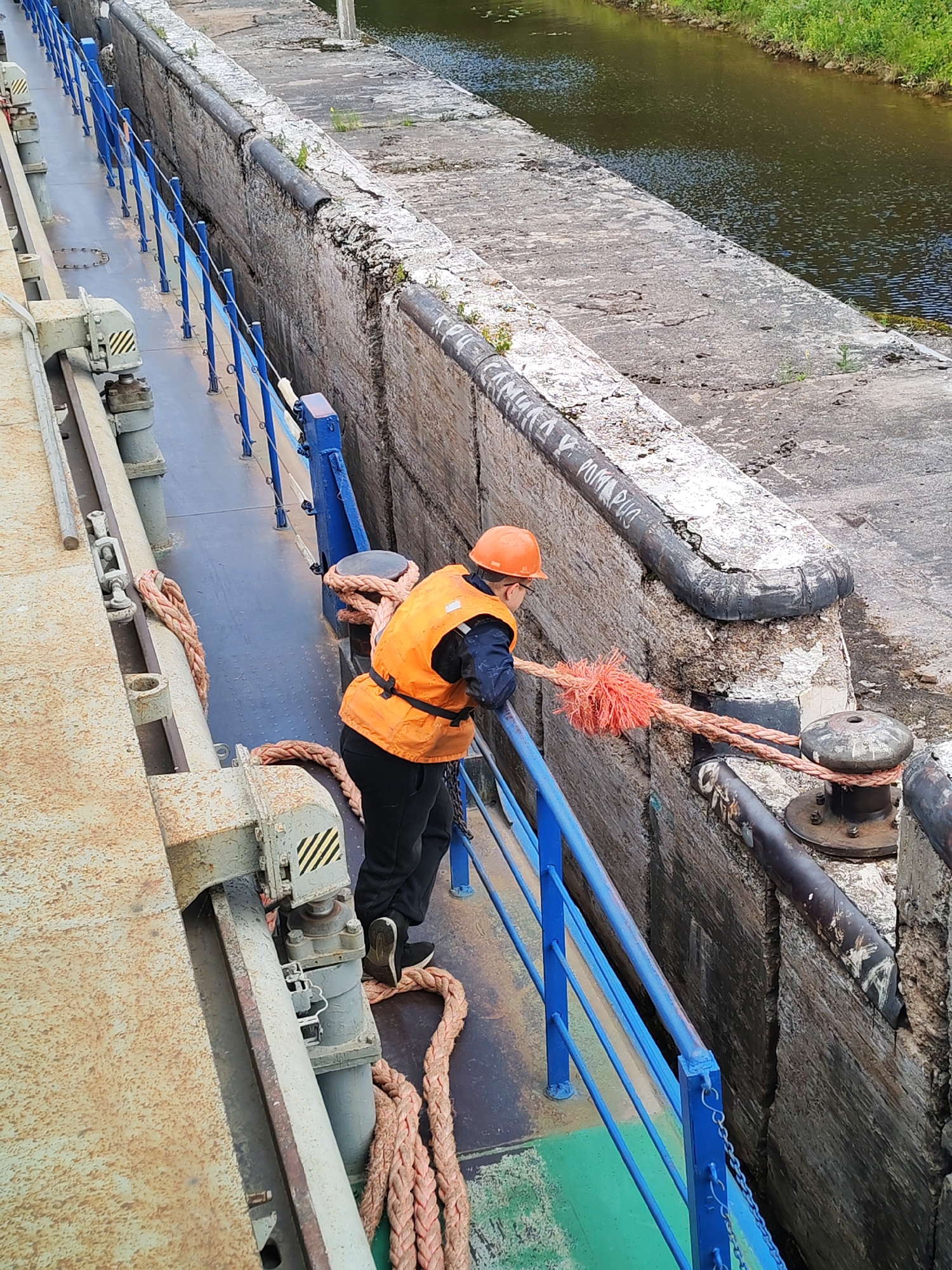 In Vytegra, Russia, a person wearing an orange hard hat and an orange safety vest is on the deck of a vessel, tending to a thick, pink rope. The rope is secured to a bollard on the concrete wall of a canal lock. The person is holding the rope taut, assisting in mooring or unmooring the vessel. The deck of the vessel is metallic and appears weathered, with a blue railing along the edge. The canal lock wall is made of rough-hewn stone and concrete, showing signs of wear and tear. The water in the canal is dark and appears calm, with green vegetation visible on the far bank. The scene suggests a routine operation within the functioning of the canal system in Vytegra.