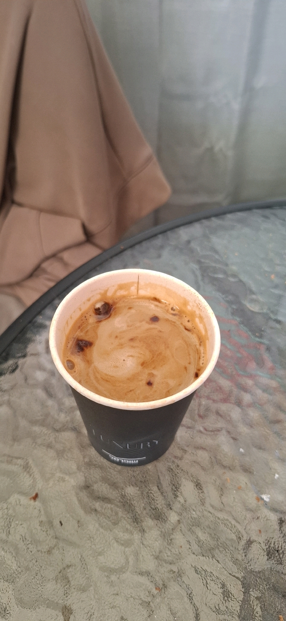 A disposable coffee cup sits on a glass table in what appears to be an outdoor seating area, possibly a cafe or residential patio. The cup, dark gray with a white rim, contains a frothy coffee with swirls of crema. The surface of the glass table is textured and reflects light, showing faint patterns. In the background, a large piece of light brown fabric, possibly an umbrella or awning, is visible, along with a blurred, light-colored surface that could be a wall or another piece of fabric.
The location is identified as Yerocham, Israel. However, there are no specific visual cues in the image to confirm this location, such as landmarks, signage, or distinct architectural features associated with Yerocham. The scene is static and shows no people or any active interactions.