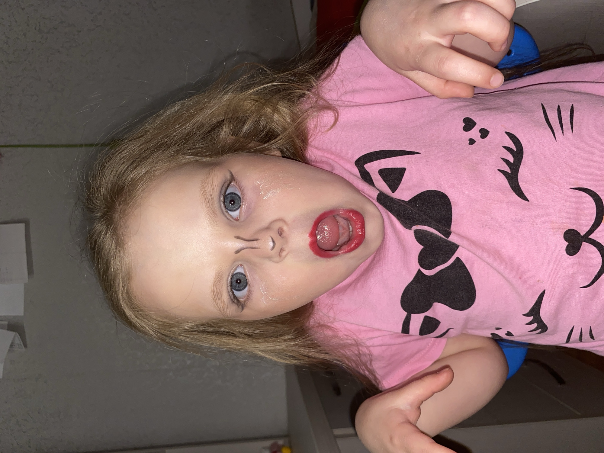A young child with light blonde hair and blue eyes is captured in an indoor setting, looking directly at the camera with a wide-eyed, open-mouthed expression. The child's lips are coated in vibrant red lipstick, applied somewhat outside the natural lip line, and faint markings resembling whiskers or lines are drawn around the nose. A slight sheen is visible on the child's skin, possibly from the makeup or natural oils.
The child is wearing a pink t-shirt adorned with black graphics, including a bow, heart shapes, and designs that resemble cat whiskers or facial features, suggesting a cartoon character or animal theme. One of the child's hands is visible in the upper right corner of the frame, near the head, while another hand is seen in the lower right, appearing to hold or rest near a blue object or garment.
The image is oriented vertically, with the child's head on the left and feet/upper torso on the right, suggesting a perspective from above, possibly with the child lying down. The background consists of a plain, light-colored wall or surface, with a faint green line visible on the left side. The lighting appears artificial, indicating an indoor environment. The overall scene suggests a playful activity, likely dress-up or experimentation with makeup, conveying a sense of childhood curiosity and mischief. No discernible text is visible in the image.