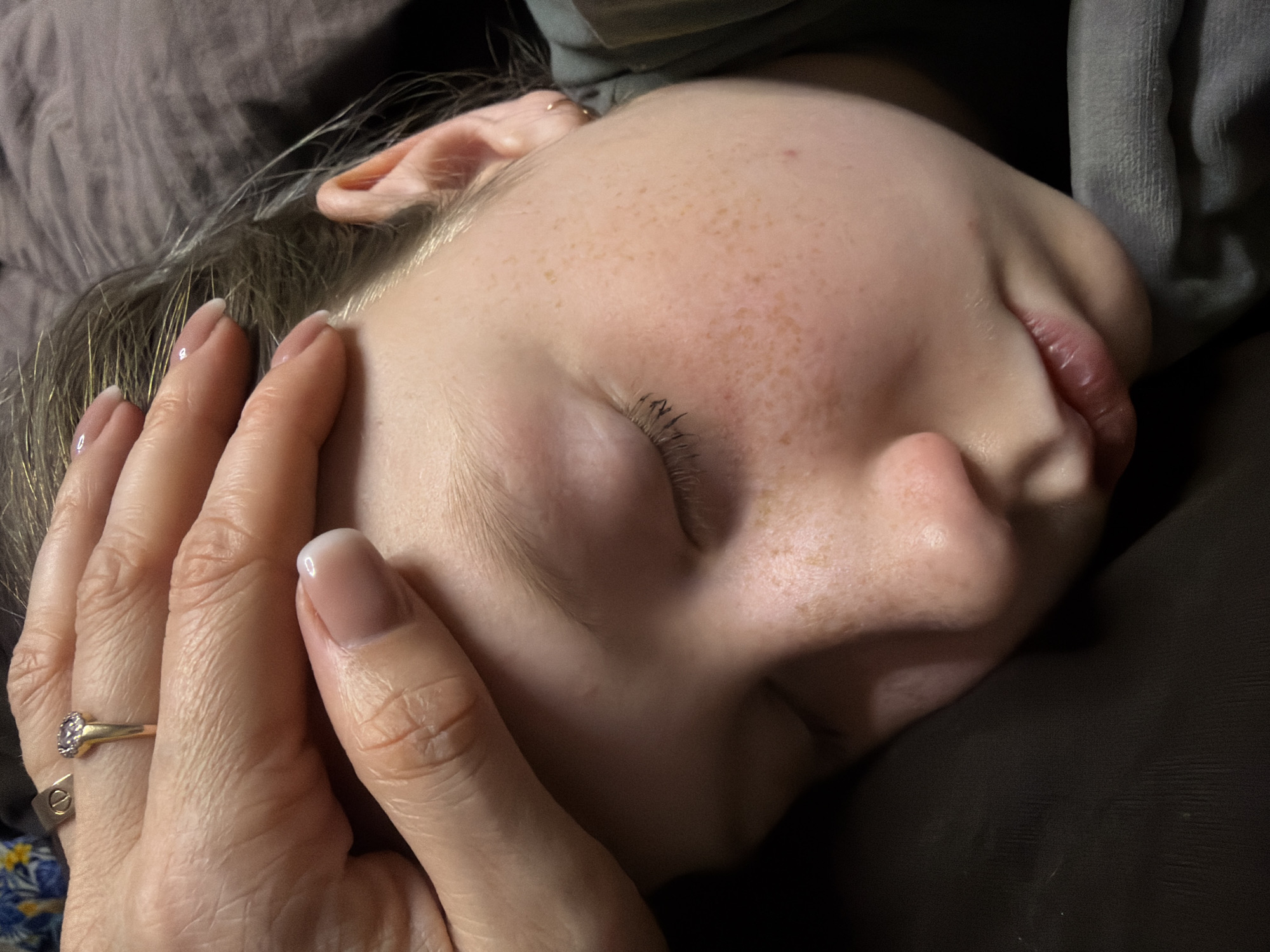 Close-up of a sleeping child's face. The child is lying on a dark surface, possibly a pillow. A hand with well-manicured nails and a gold ring is touching the child's cheek. The child has fair skin with freckles and closed eyes. There is no information available to confirm this scene is in Dubovaya, Russia.