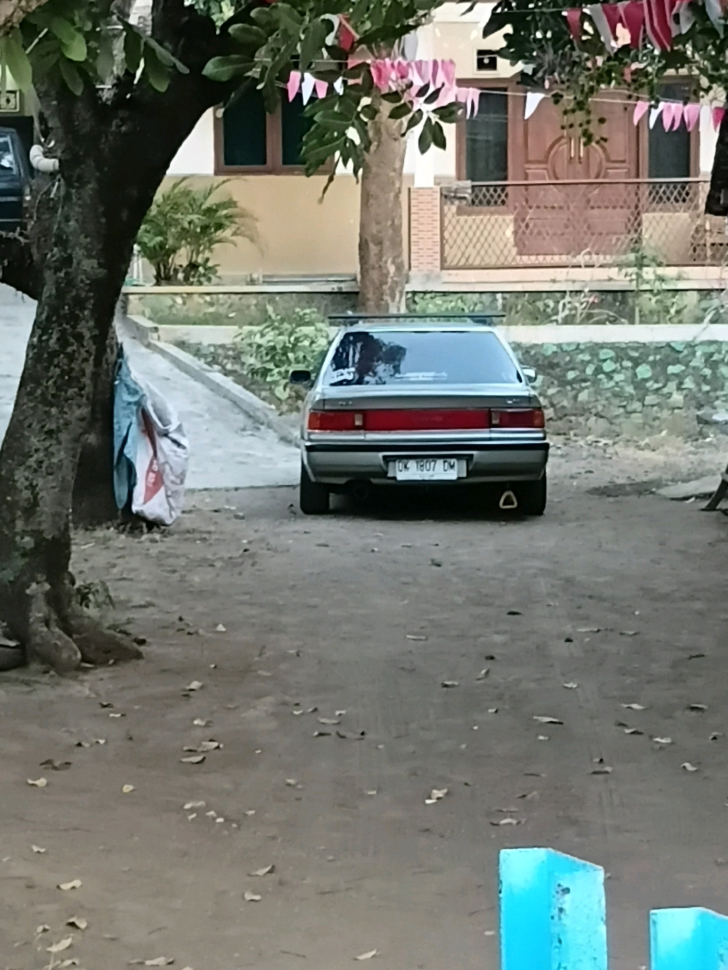 A silver sedan is parked on a dirt road in front of a building. The car's license plate reads "DK 1807 DM". There is a tree with a large trunk to the left of the car, and behind the car, a building with a brown door and windows. Small flags are strung between trees in the background. A blue structure is visible in the lower right corner of the image. The scene is likely in Kebonsari, Indonesia, given the Indonesian license plate.