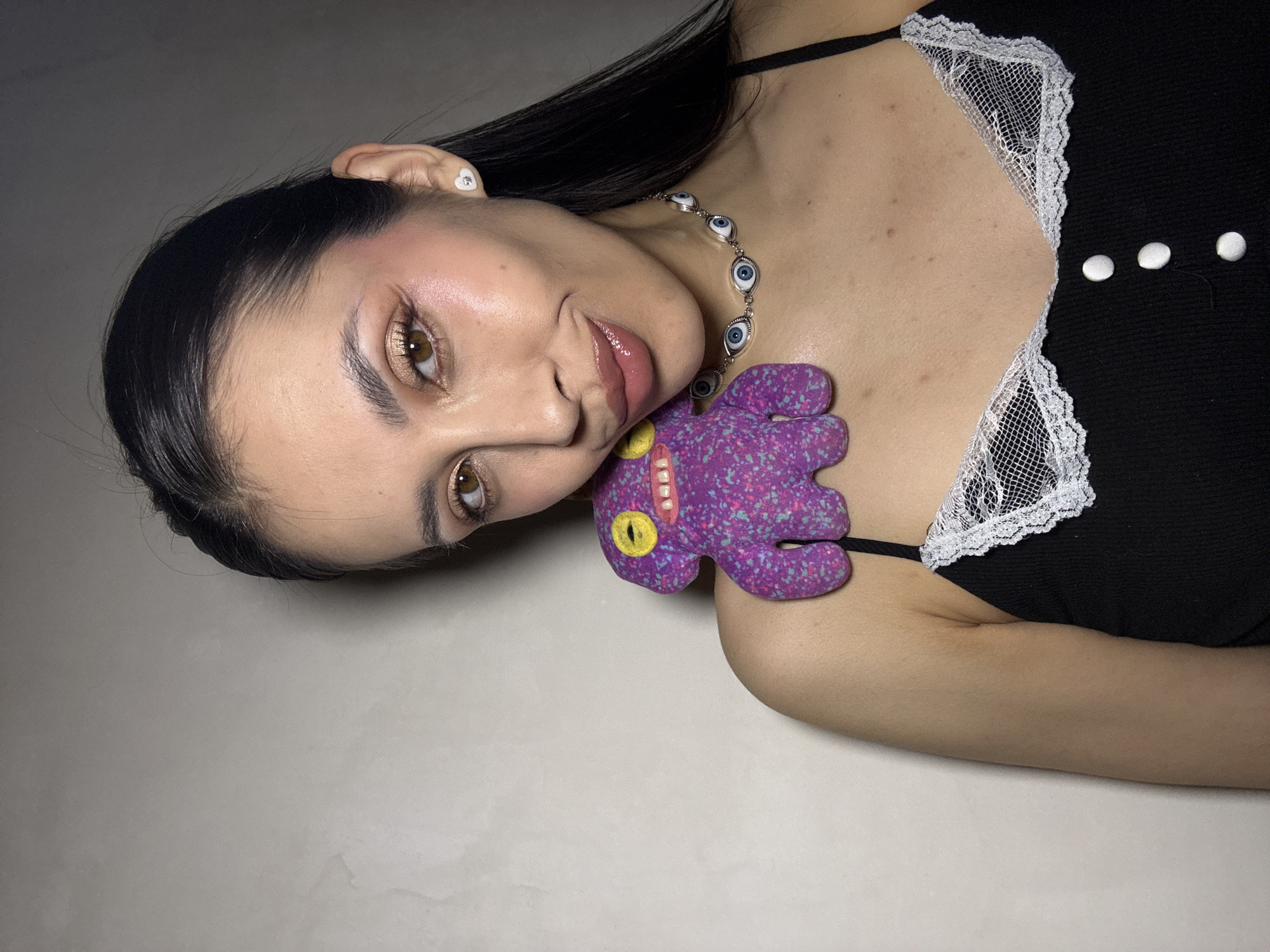 A woman is lying down, partially rotated towards the camera. Her dark hair is neatly styled, and she wears eye makeup. She is wearing a black top with white lace trim and three white buttons. A necklace with stylized eye charms adorns her neck. A plush purple octopus with yellow eyes and a pink mouth is positioned against her neck. The background is a plain, light-colored surface. There is no indication of the city of Votkinsk, Russia.