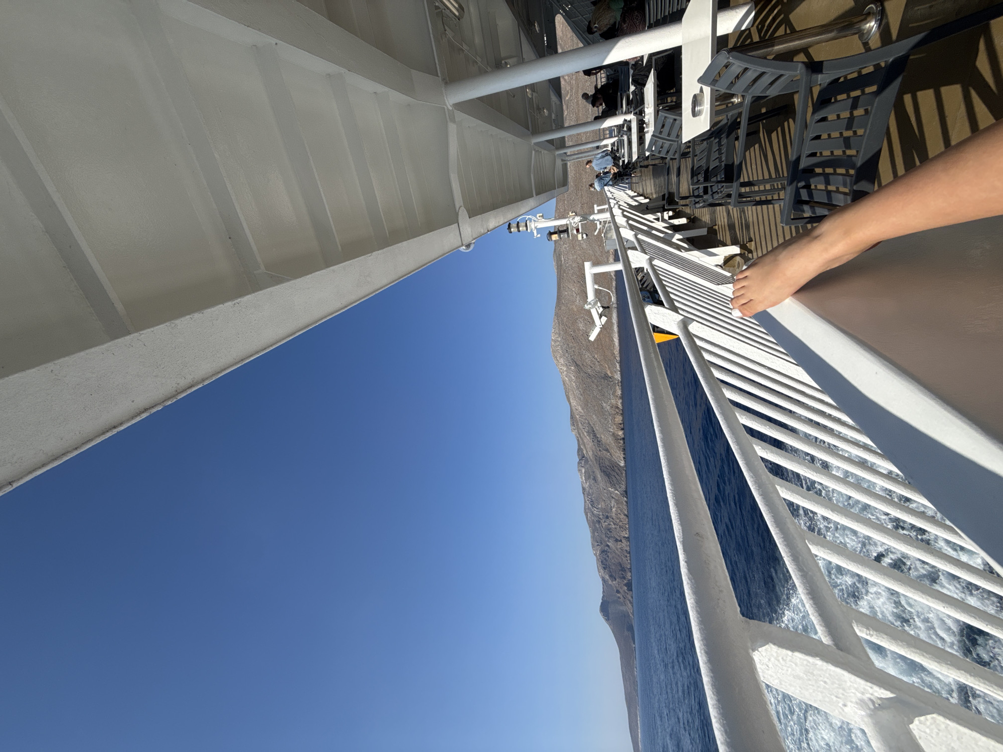 The image shows a view from the deck of a ferry sailing past a mountainous coastline. The sky is a clear, bright blue, suggesting a sunny day. The sea is a deep blue, with whitecaps indicating moderate waves. The ferry has a white railing along its deck, which dominates the foreground and mid-ground on the right side of the image.
In the background, a rocky, arid mountain range rises from the sea, its slopes catching the sunlight. The scale of the ferry and the surrounding landscape suggests it is a passenger ferry. On the upper deck of the ferry, several figures are visible, some sitting at tables, and one person's bare leg is prominently placed in the foreground, resting on the railing. This placement suggests a relaxed and possibly leisurely atmosphere.
The scene is set during the daytime, likely late morning or early afternoon, given the bright sunlight and clear skies. The location is identified as Livadia, Greece, which aligns with the arid, mountainous coastal scenery. There is no visible text in the image. The overall mood conveyed is one of travel, relaxation, and enjoying a scenic journey.
