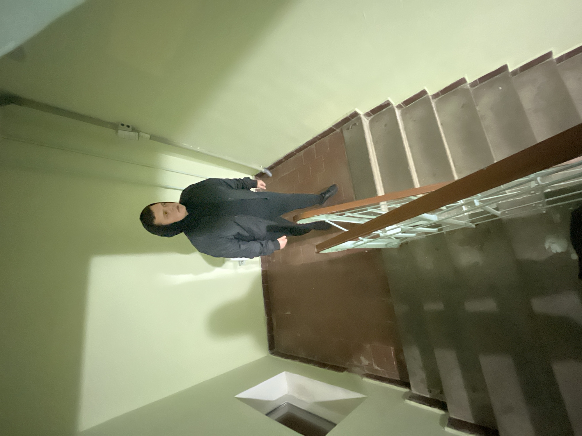 A man in a black jacket and pants stands on a landing of a staircase in an indoor space. The walls are a pale green, and the staircase has brown treads and white risers. A wooden handrail with a metal railing is present. The space appears to be a stairwell in a building. A window opening to the floor below is visible. There is no information to confirm that this image was taken in the city of Klin, Russia.
