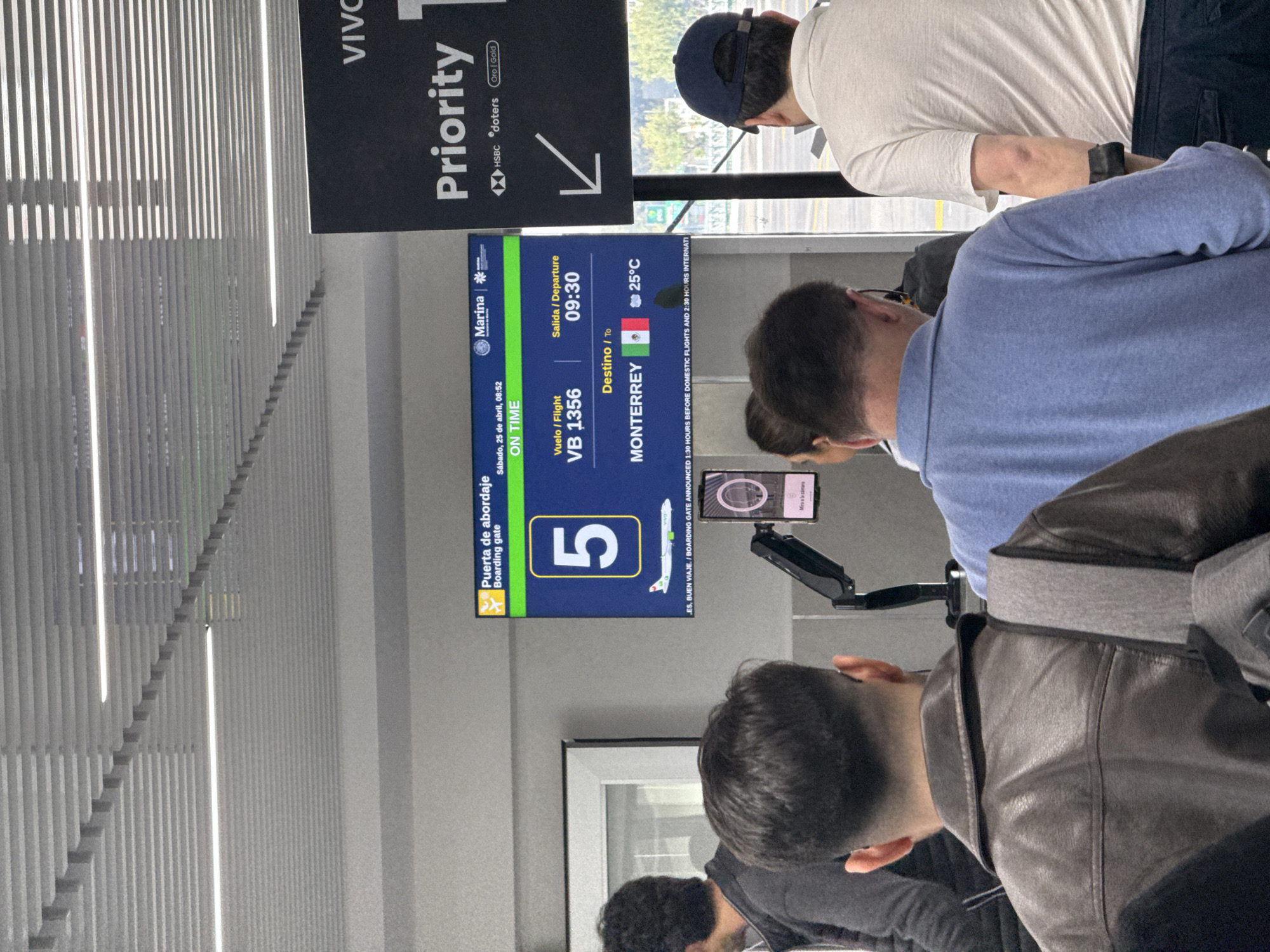 The image shows a group of people at an airport, likely in Mexico City, Mexico. A digital sign displays flight information, including the boarding gate number 5, flight number VB 1356, destination as Monterrey, departure time of 09:30, and a temperature of 25°C. The sign also states that boarding gate announcements are made 1.5 hours before domestic flights and 2.5 hours before international flights. A "Priority" sign is also visible. Several people are in the foreground, partially obscuring the view of the sign. One person is holding a smartphone pointed towards the sign.