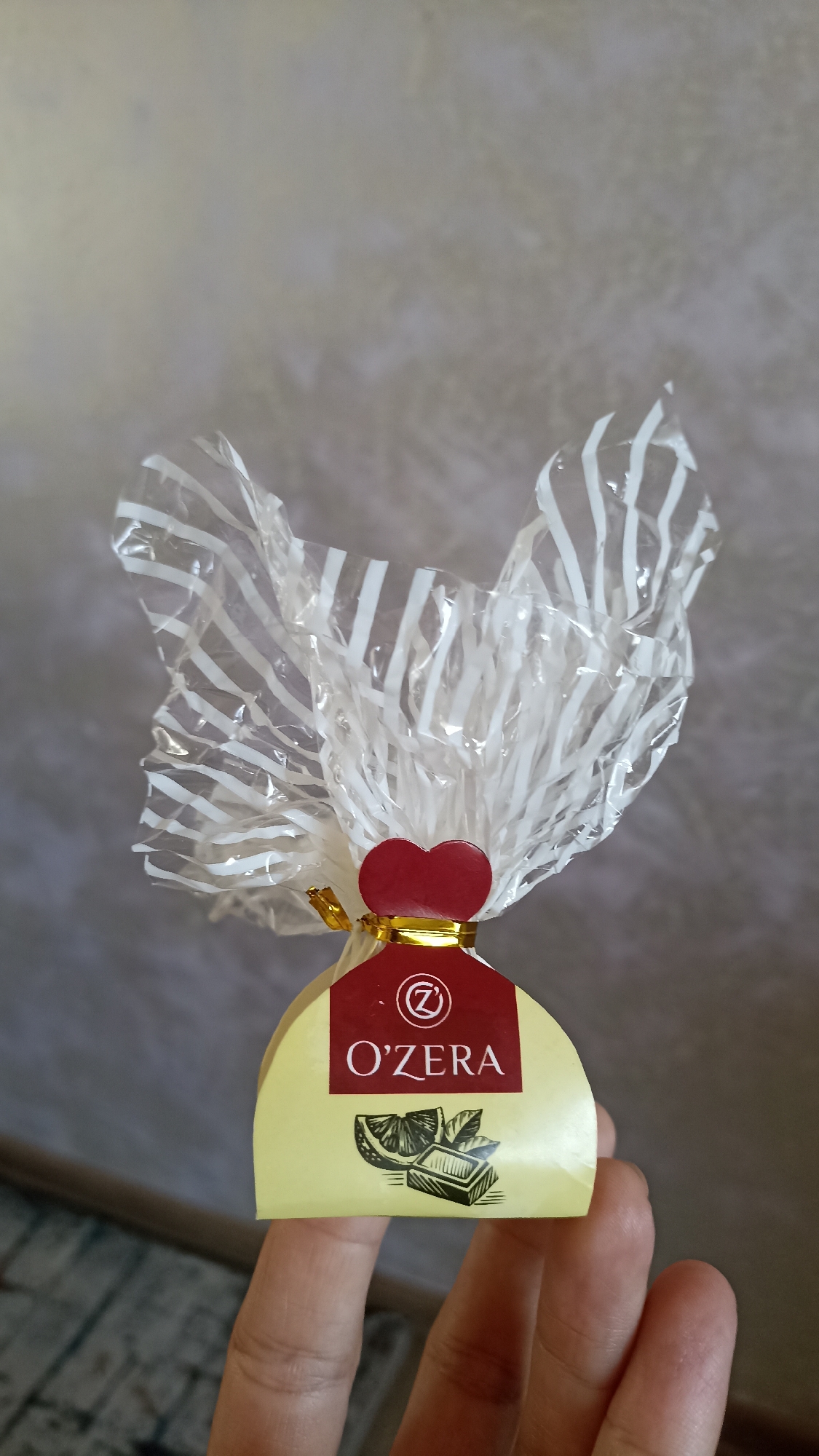 A person's hand is holding a piece of candy. The candy is wrapped in translucent, crinkled plastic with white stripes. A red heart-shaped charm is tied around the neck of the candy with a gold ribbon. The candy itself is a pale yellow, semi-circular shape with a label. The label is dark red with the text "O'ZERRA" and a stylized "Z" in a circle. Below the text are illustrations of a lemon slice and a piece of chocolate. The background is a textured, light-colored wall. The image does not contain any information about the city of Ak-Suu, Kyrgyzstan.