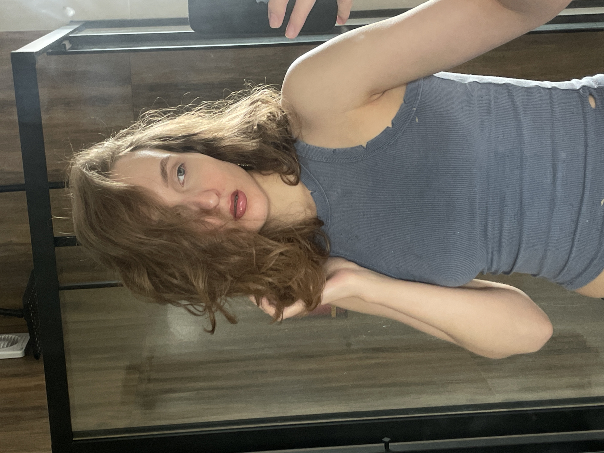 A young, light-skinned individual with long, brown, curly hair and wearing a gray tank top is depicted in an indoor setting. The person is lying down, with their head tilted towards the camera. The background appears to be a floor with a wood-like pattern, and there is a black metal frame of a structure (possibly a table or furniture) partially visible. The location of the image is Unknown, Unknown.