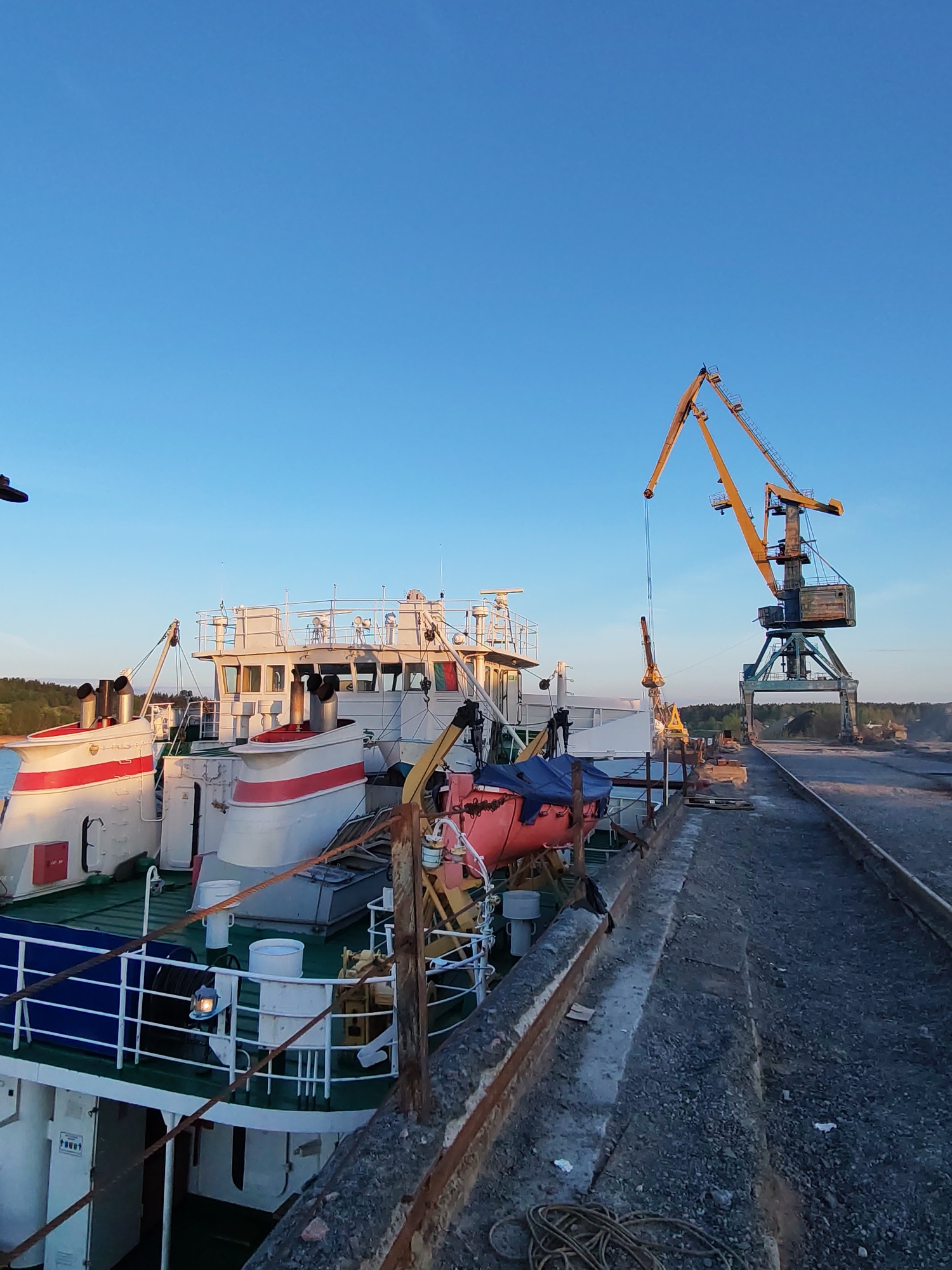 The image shows a ship docked at a pier in Tver, Russia. The ship is white with red and blue trim. It has multiple decks and a lifeboat. Adjacent to the ship is a concrete pier with some gravel and debris. Two large yellow cranes stand on the pier near the ship. The background reveals a clear blue sky. The scene appears to be at dusk or dawn due to the lighting conditions.