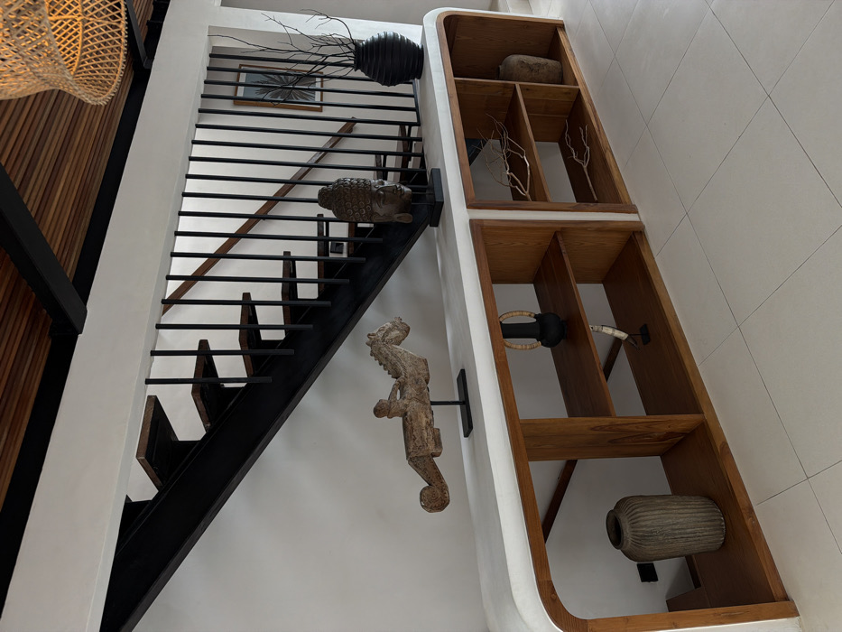 A modern interior showcases a black staircase with a horizontal railing design, accented by a wall-mounted Buddha head and a seahorse statue. The staircase ascends adjacent to a built-in wooden shelving unit displaying decorative items such as rocks, branches, a light fixture, and a textured vase. The color palette is predominantly white, contrasted by the black stairs and wooden elements.