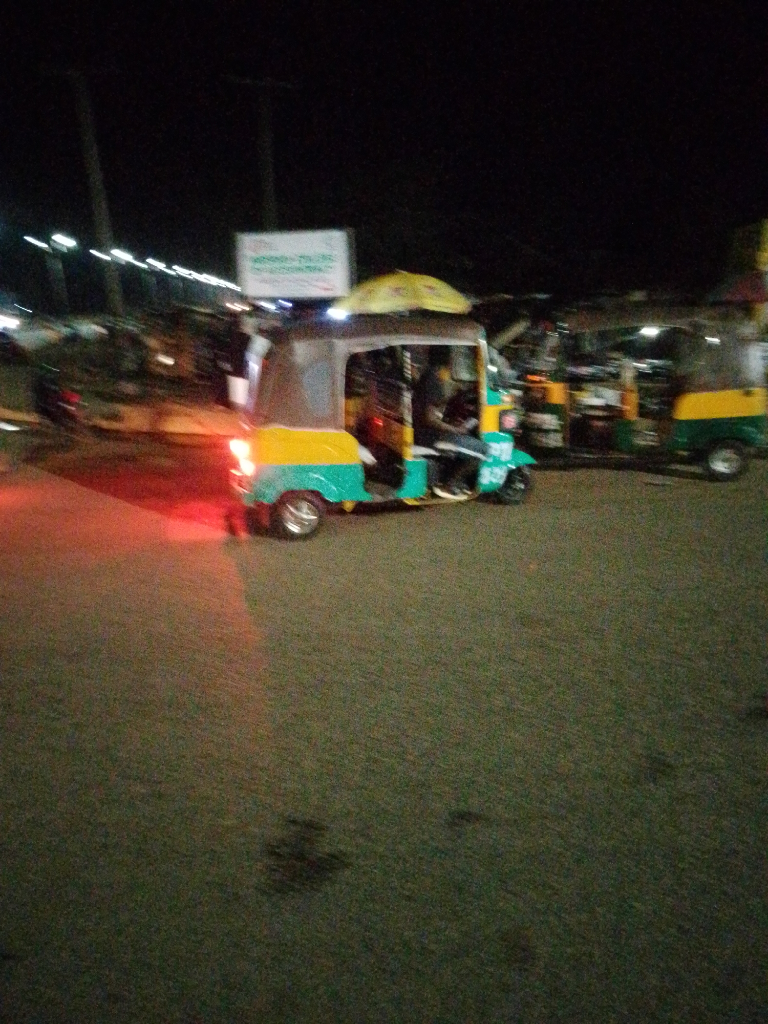 An auto-rickshaw with a yellow and teal canopy is parked on a paved surface at night. The vehicle is illuminated by red brake lights. The background is dark, with some blurred lights visible, suggesting it is a street or an area with some activity. The ground appears to be asphalt. The overall image quality is poor due to low light conditions and motion blur. This scene is located in Bukuru, Nigeria.