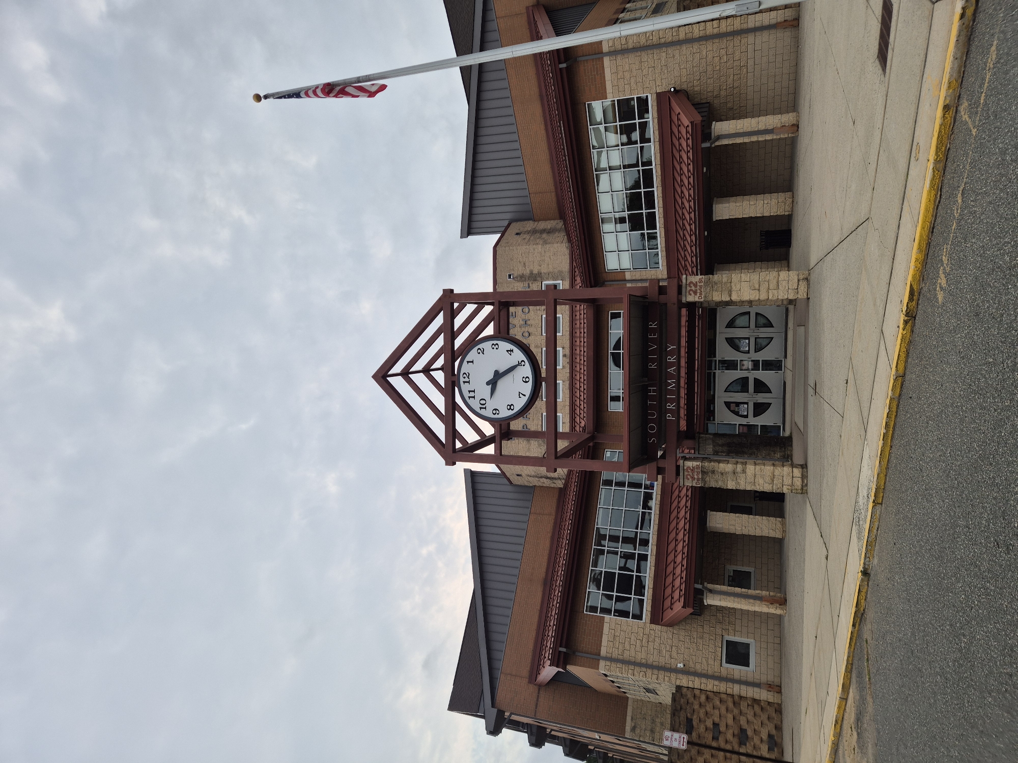 The image shows the front entrance of South River Primary School, located in South River, New Jersey, United States of America. The building features a brick facade with stone accents and a prominent clock above the main entrance. An American flag flies from a flagpole to the right of the building. The sky is overcast with scattered clouds. A sidewalk and street are visible in the foreground. There are no people in the image.