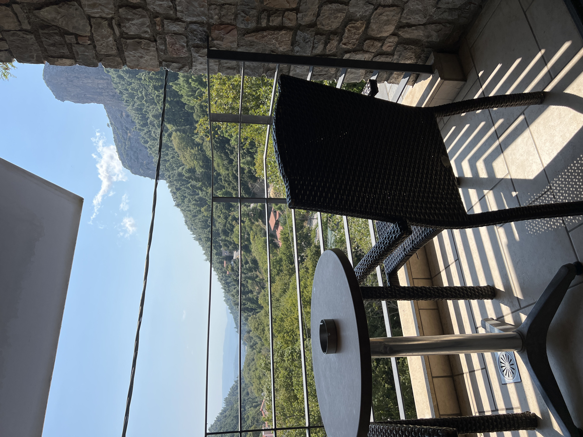 The image depicts a view from a balcony in Στενή Δίρφυος, Greece. In the foreground, a round table with an ashtray sits on a tiled balcony floor, illuminated by strong sunlight casting distinct shadows from a chair. The chair, made of dark, woven material, is positioned to the right of the table. A metal railing separates the balcony from the scenic landscape beyond. The background reveals a lush, green valley dotted with buildings, leading to distant mountains under a clear blue sky with scattered clouds. The architectural style of the buildings suggests a traditional Greek village setting, and the rugged mountain terrain is characteristic of the Greek mainland, specifically the island of Evia where Στενή Δίρφυος is located, known for its natural beauty and hiking trails. The stone wall on the right edge of the frame indicates the balcony's construction.