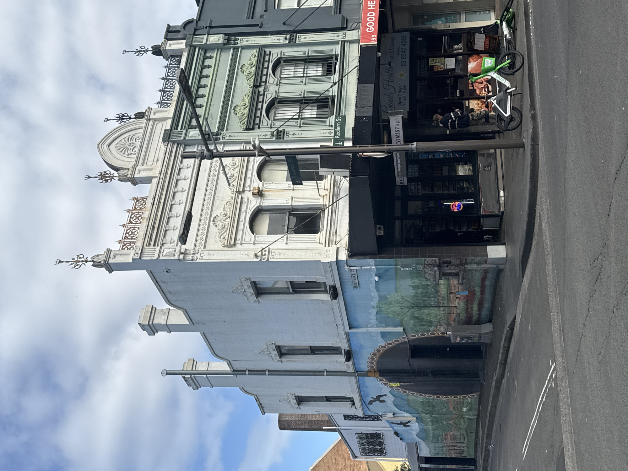 The image captures a street scene in Glebe, Australia, featuring historic buildings with ornate architectural details. The main building on the left is painted white and adorned with a colorful mural depicting a landscape with birds, trees, and figures. The building to its right is painted a light green and exhibits decorative stonework and arched windows. A street sign indicates "Renwick St" on the left building and "Redfern St" on the green building. A street lamp stands in the foreground, positioned between the two buildings. In front of the shops, a person is visible standing near the entrance of "Good Health Fine Massage". A white and green Lime electric scooter is parked on the sidewalk, and another electric scooter is lying on its side nearby. The street in front of the buildings is paved, with double white lines indicating a pedestrian crossing. The sky above is partly cloudy.