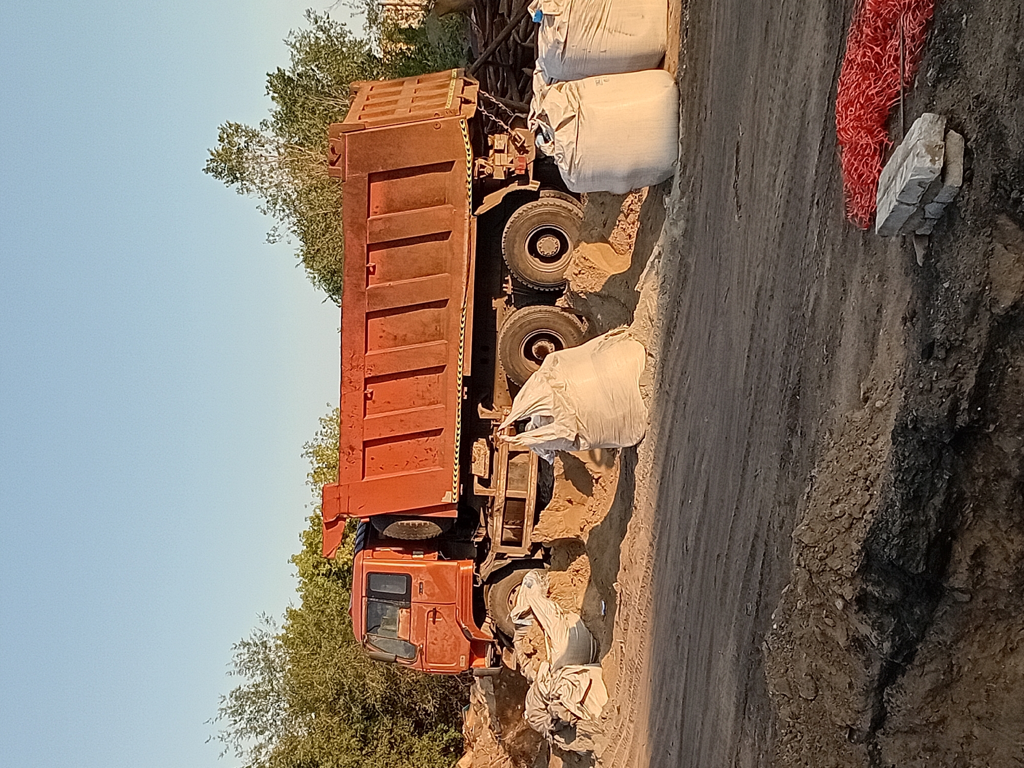 A large, orange dump truck is partially submerged in sand at a construction site. The truck is positioned at an angle, with its rear wheels dug into the sand. Several large white bags, likely filled with sand or construction materials, are also present in the foreground. In the background, there are trees and a clear blue sky. The ground is a mix of dirt and sand, with tire tracks visible. A pile of red netting and some bricks are in the lower right corner of the image. The overall scene suggests ongoing construction or demolition work. This scene is located in the city of Semey, Kazakhstan.