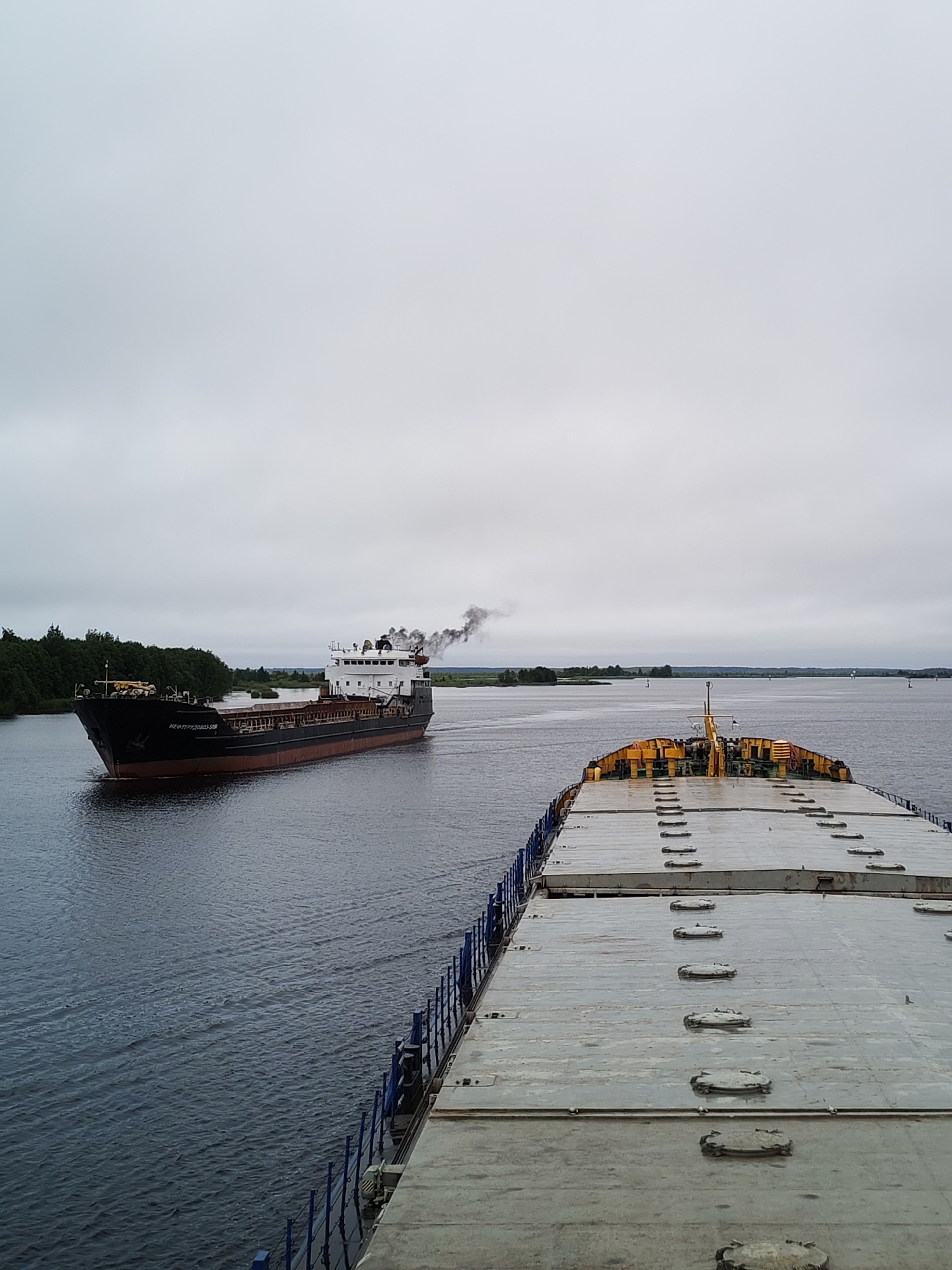 The image depicts a wide, calm river under an overcast sky. In the foreground, the deck of a large, flat-topped barge or cargo ship is visible, extending towards the viewer. This vessel is loaded with what appear to be large, rectangular containers or cargo slabs, painted a light grey. The barge has blue railings along its edge.
Further down the river, a black and red cargo ship is sailing from left to right. This ship is emitting a plume of dark smoke from its funnel. The ship has visible markings, including the text "НЕФТЕПЕРЕРАБ.ЗАВОД" in white lettering on its hull. The riverbanks are lined with dense green trees, suggesting a rural or natural setting. The overall atmosphere is subdued due to the cloudy weather. The location is identified as the city of Novokemskii, Russia.