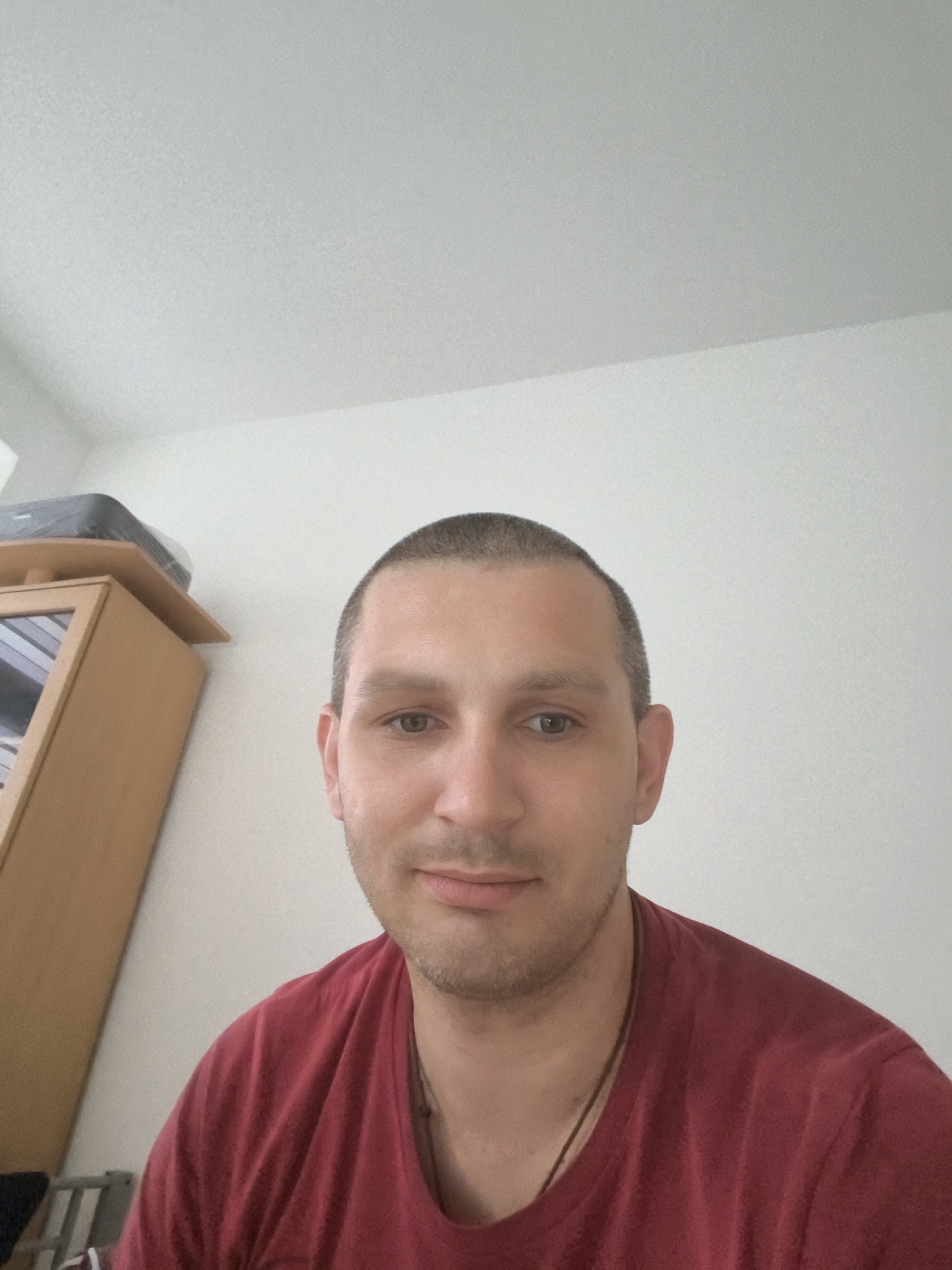The image shows a close-up, waist-up portrait of a man with a shaved head and light stubble, wearing a red t-shirt. He is looking directly at the camera with a neutral expression. In the background, to the left of the man, is a wooden cabinet with a glass-fronted door, and on top of it, a black electronic device wrapped in plastic. The wall behind the man appears to be white and textured. The lighting is natural and diffused, suggesting an indoor setting. The image does not provide any visual cues or information that would allow for a determination of its location within the city of Wolgast, Germany.