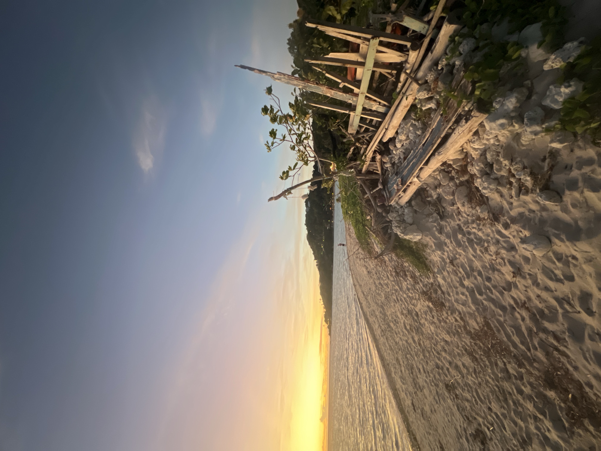A sandy beach meets calm ocean waters under a gradient sky, transitioning from a pale blue at the top to a warm yellow and orange glow near the horizon. The sun appears to be setting or rising, casting a soft light across the scene. On the shore, a pile of weathered wooden debris is scattered amongst rocks and vegetation, suggesting a natural or possibly man-made disturbance. A lone figure stands near the water's edge in the distance, looking out towards the sea. The sand is textured with ripples and scattered debris. The overall mood is tranquil and serene, evoking a sense of a peaceful moment at a tropical beach. No text is visible in the image.