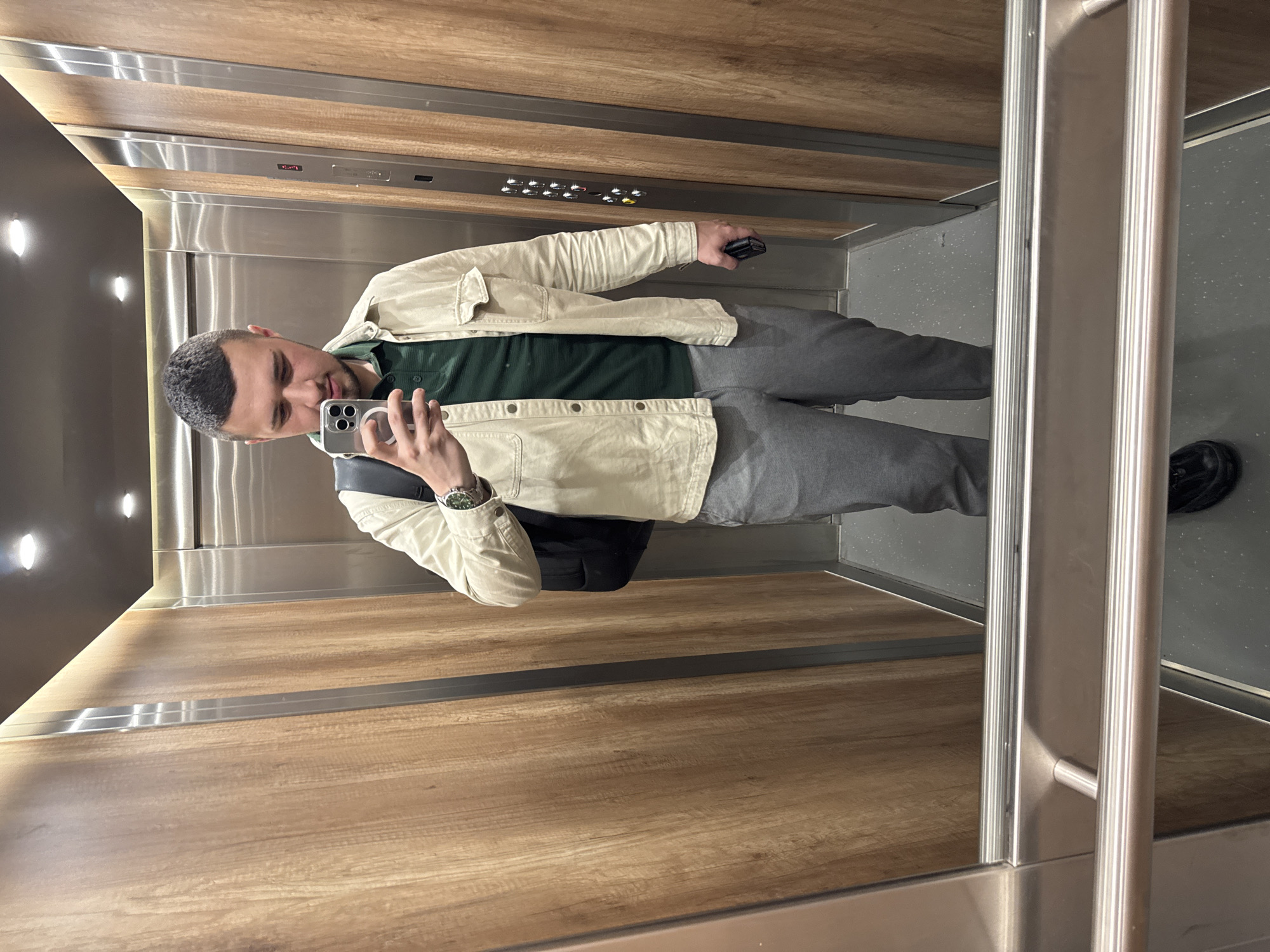A man is captured taking a mirror selfie inside a modern elevator, located in Sofia, Bulgaria. The image is oriented horizontally, showing the man's reflection as he holds up an iPhone with a multi-lens camera setup. He is dressed casually in a light cream-colored, long-sleeved jacket worn over a dark green t-shirt, and grey sweatpants. A black backpack is visible on his left shoulder, and he wears a watch with a green bezel and silver strap on his left wrist. His expression is neutral and relaxed, directed towards his phone screen.
The elevator interior features a contemporary design with wood-paneled walls in the lower half and reflective stainless steel panels above. A control panel with multiple buttons and indicator lights is visible on the stainless steel wall, reflected from the opposite side of the elevator. The floor is a speckled grey. Two oval-shaped light fixtures illuminate the left wall, and a stainless steel handrail or frame is prominent on the right side of the reflected space. The scene depicts an everyday moment inside what appears to be a well-maintained building.