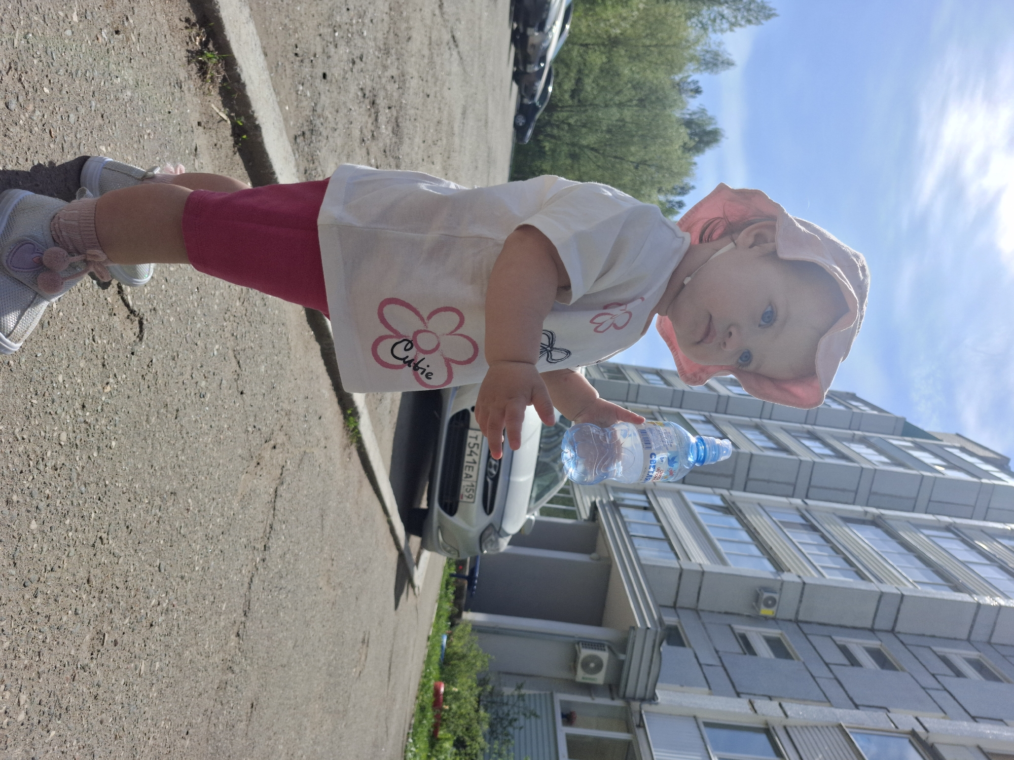 A toddler in a pink sun hat, white t-shirt with a floral design, and pink shorts stands outdoors, holding a clear plastic water bottle. The child is on asphalt, next to a curb. A silver car is parked nearby, with a license plate that appears to read "T541EA159." A multi-story residential building is in the background with multiple windows and AC units. The sky is blue with bright sunlight. There is a row of trees and another vehicle in the distance. This image appears to have been taken in Chaikovsky, Russia, based on the license plate.