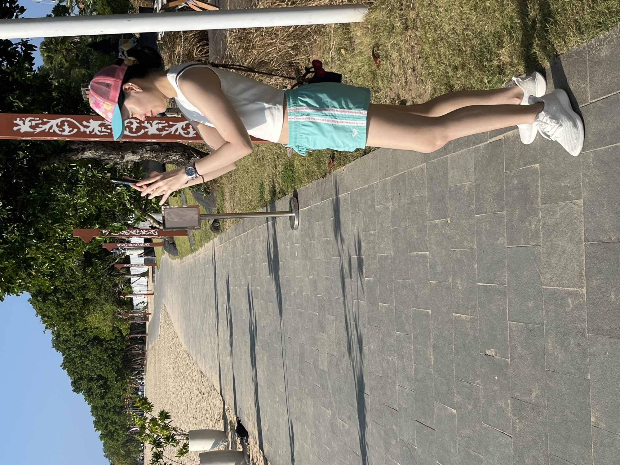 A woman is standing on a paved walkway next to a sandy beach in Kuta Selatan, Indonesia. She is wearing a white crop top, turquoise shorts with pink stripes, a pink and blue baseball cap, and white sneakers. She is holding a smartphone and appears to be taking a picture or video. The walkway is made of gray stone tiles and is lined with trees and decorative poles on the right. The beach to the left is sandy, and beyond it are more trees and structures. The sky is clear and blue. There are shadows cast on the walkway from the trees and poles.