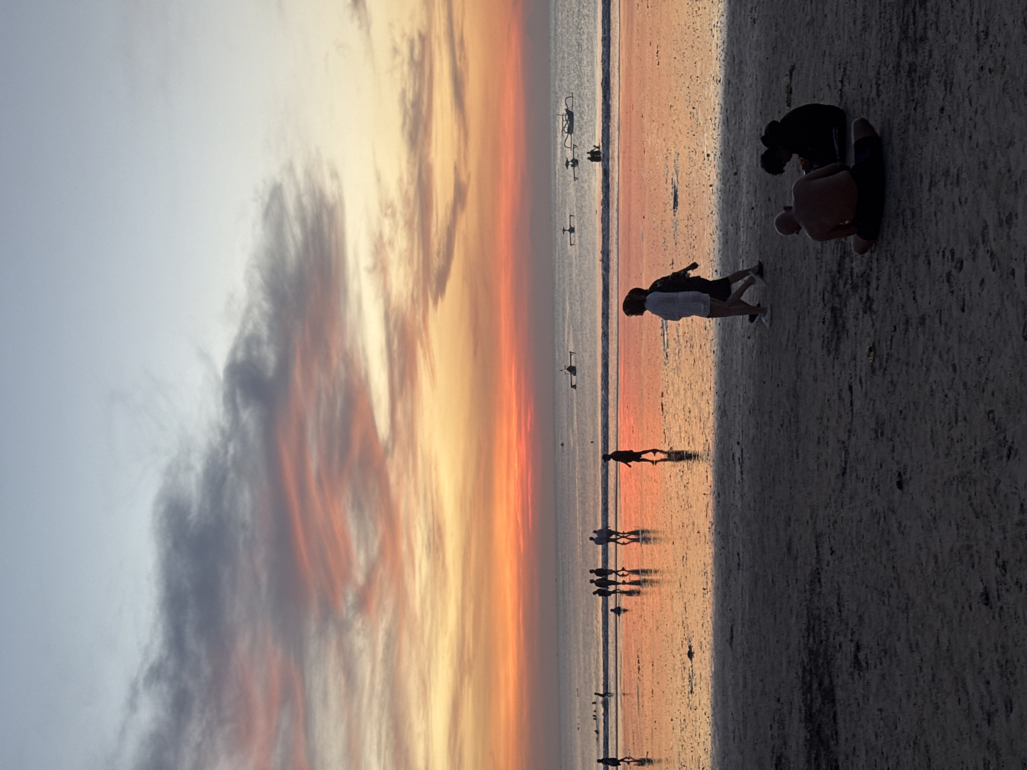 A long shot captures the sky filled with orange and gray hues during sunset. Below, on a beach, silhouettes of people stroll along the shoreline. In the foreground, two people appear to be tending to someone on the sand. The location is the beach in Kuta Selatan, Indonesia.