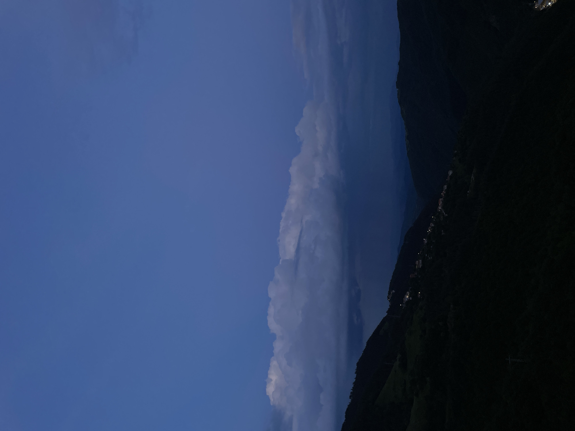 The image depicts a landscape at dusk in the city of Pechinska, Bulgaria. The sky occupies most of the left side, with a gradient from light blue to darker blue, suggesting the sun has set or is setting. A large bank of cumulonimbus clouds runs vertically through the center of the frame. Dark, wooded mountains rise up on the right side. There are small light orbs that are houses on the right, likely situated on the mountainsides. A telecommunication pole is seen on the bottom right corner. The overall environment appears rural, at dusk.