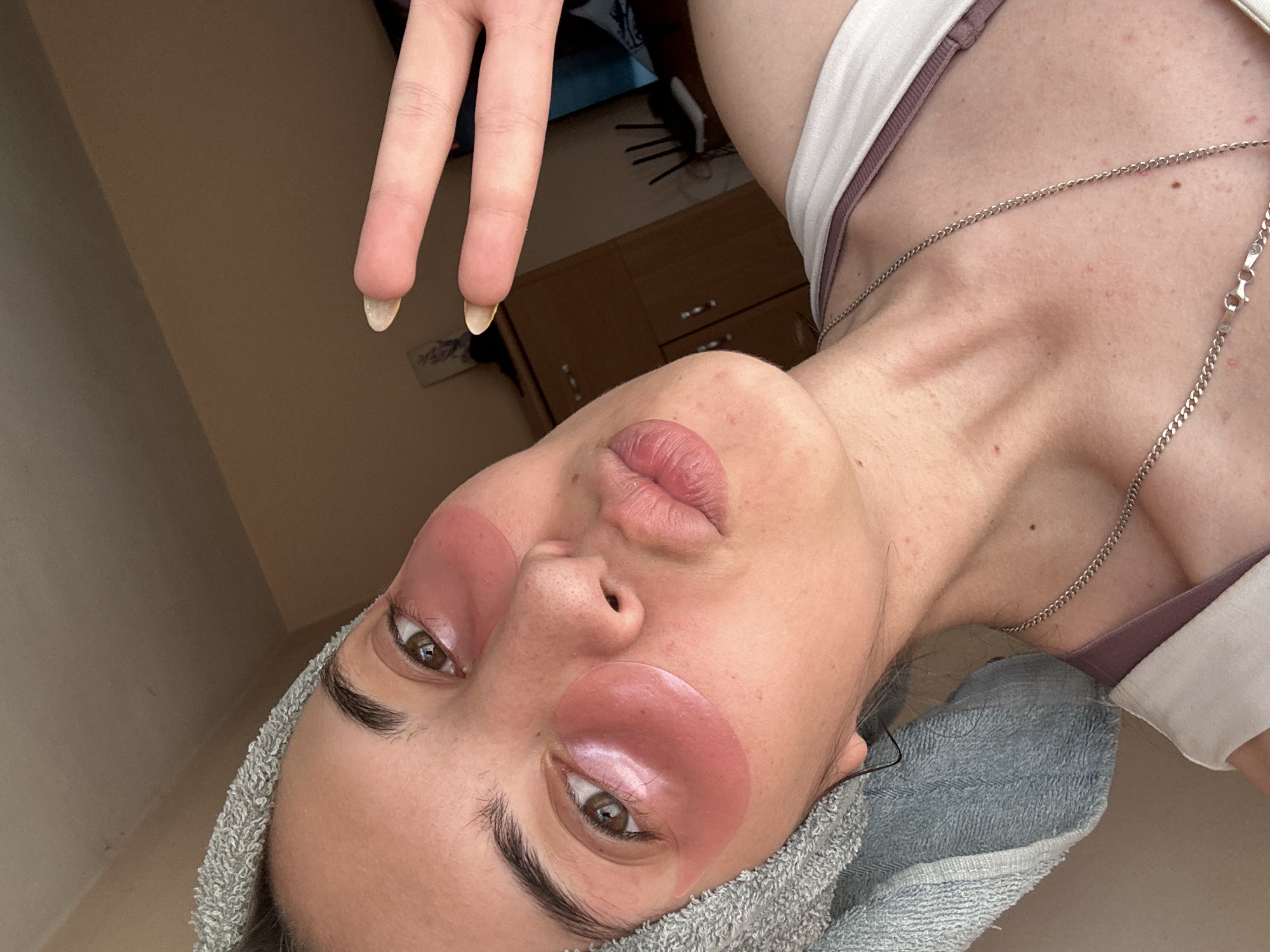 A young adult female is depicted in a close-up selfie. She has fair skin, brown eyes, and is wearing pink eye masks. She has a gray towel wrapped around her head. A silver necklace is visible around her neck. Her lips are slightly pursed. Two fingers of her left hand are visible in the upper left corner of the image. The background appears to be an interior setting with neutral-toned walls and furniture. The image does not provide any specific indicators to confirm the location of Sirakovo, Bulgaria. The scene depicts a personal grooming or self-care activity.