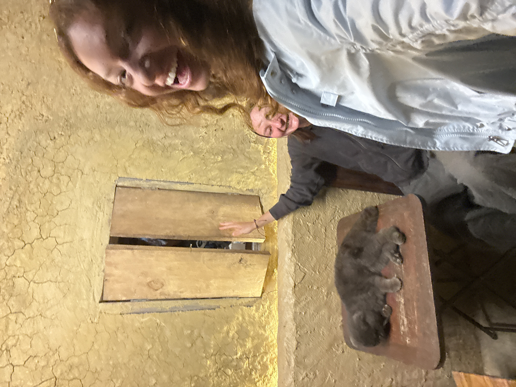 Two smiling women are visible in the frame, one with red hair. A gray cat sleeps on a red-brown surface. One woman's hand touches wooden window shutters set into a rough, textured yellow wall. The scene may be located in Lũng Cú Commune, Vietnam.