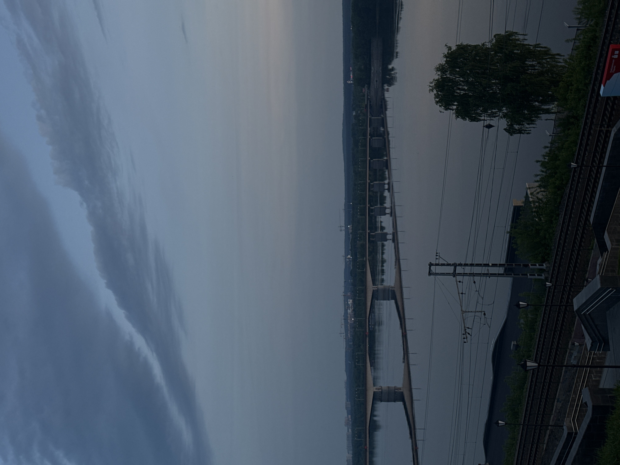 The image shows a twilight scene of the Kama River and its surroundings in Perm, Russia. A long bridge spans the river, supported by regularly spaced concrete pillars. On the right side of the image, a railway track runs parallel to the river's edge, complete with overhead power lines and support structures. Vegetation grows along the side of the railway. The sky is overcast with distinct cloud formations.