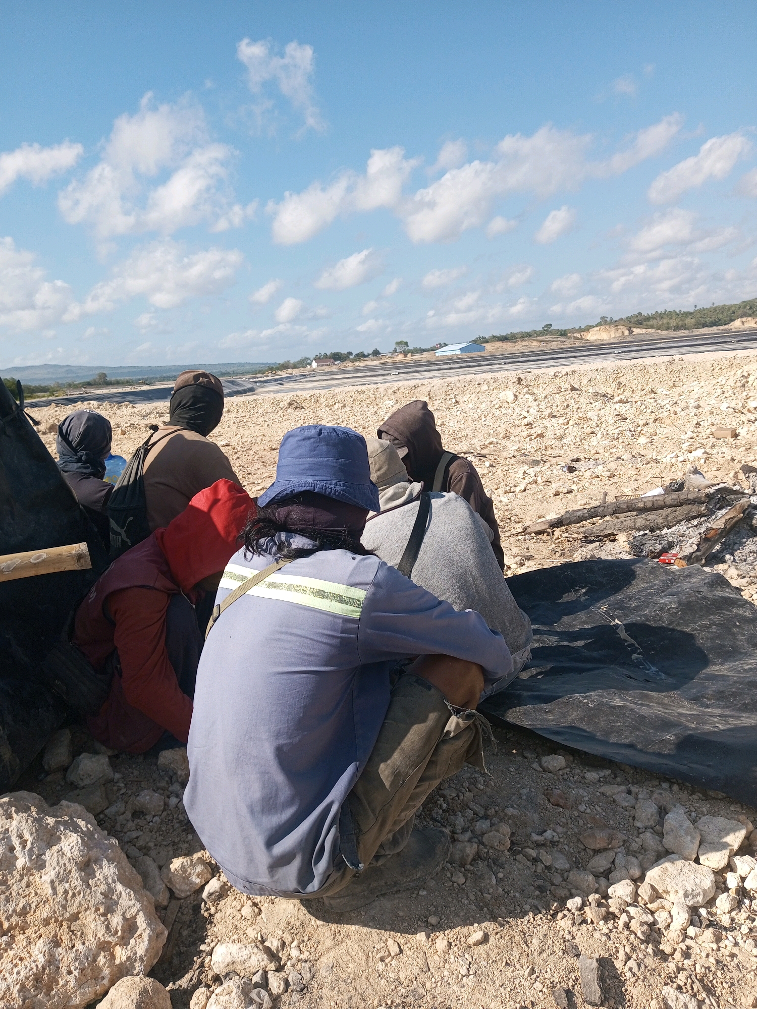 The image depicts a group of individuals, seen from behind, sitting on the ground in a desolate, rocky area. They appear to be in a construction or industrial zone, with evidence of ongoing work in the background, including what looks like a road or pathway and some large tarps. The individuals are dressed in casual clothing, some wearing hats and hoods, suggesting protection from the sun or elements. The environment is dry and arid, with scattered rocks and sparse vegetation. The sky is blue with scattered clouds. The overall impression is one of a work site or a temporary encampment in a harsh outdoor setting. While the image itself does not contain specific identifiers for Matasio, Indonesia, the scene is consistent with an industrial or construction area that might be found in such a location.