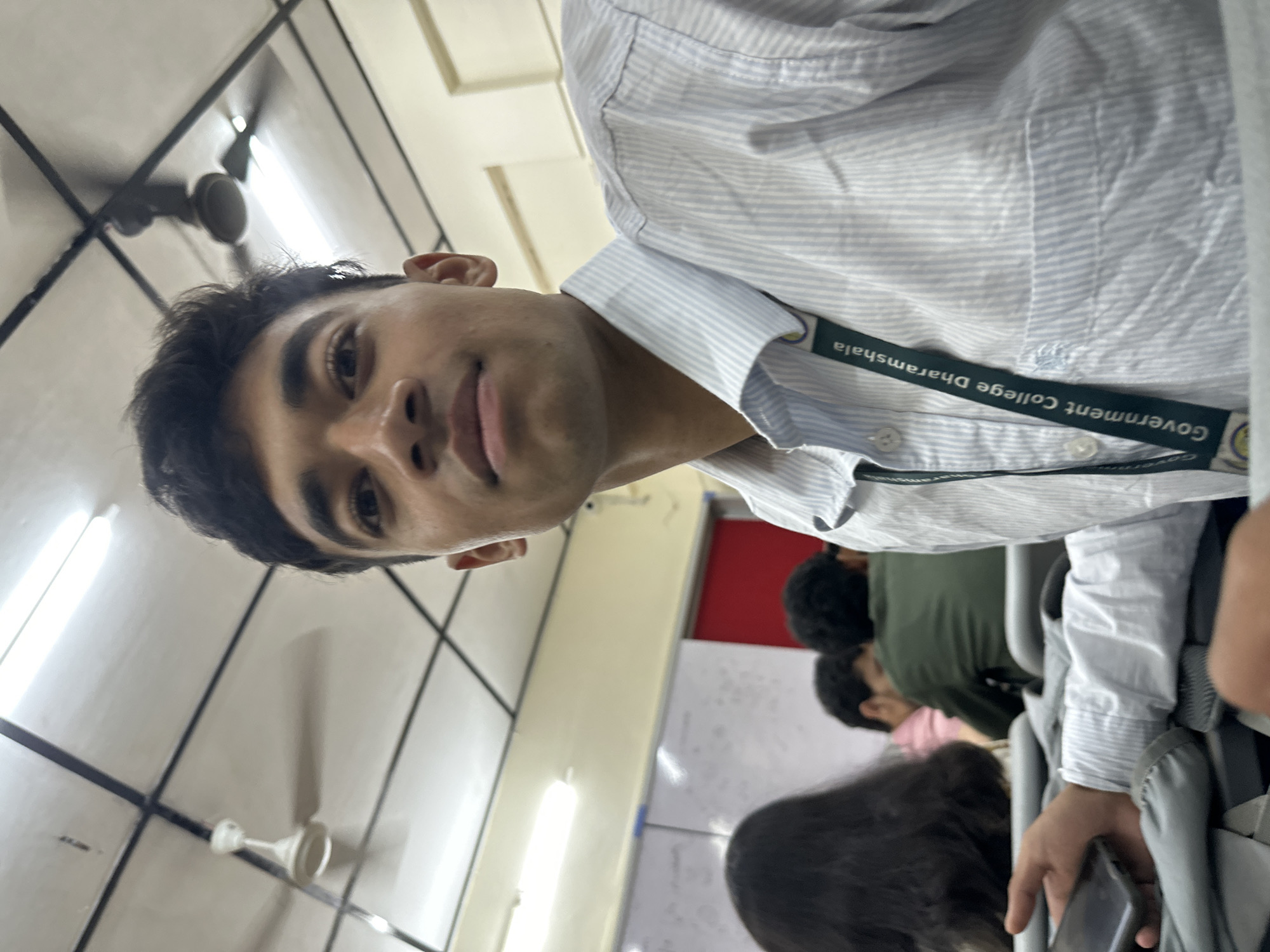 A young South Asian man wearing a light blue and white striped collared shirt is captured in a selfie taken indoors, likely in a classroom or lecture hall. He has dark, wavy hair and is looking directly at the camera with a slight smile. A green lanyard with text that reads "Government College Dharamshala" is visible around his neck, tucked into his shirt. His right hand is visible holding a black smartphone. In the background, other students are seated at desks, some with their backs to the camera. The environment appears to be a classroom with fluorescent lighting and a ceiling fan. A whiteboard with writing is partially visible on the left. The overall scene suggests a casual, everyday moment in an educational setting.