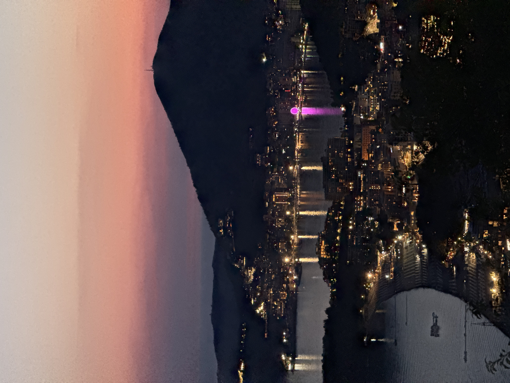 The image depicts a twilight view of the city of Podbadac, Montenegro, from an elevated vantage point. The sky transitions from a soft pink and orange hue at the top to a deep purple and dark blue closer to the horizon. A silhouetted mountain dominates the upper right portion of the frame, with a small communication tower visible on its peak. Below the mountain, the city lights of Podbadac twinkle along the coastline and hillsides, reflecting in the dark water of the bay. A Ferris wheel, illuminated in pink, is a prominent feature in the city's illuminated skyline. The foreground consists of dark, silhouetted foliage, obscuring some of the lower parts of the scene. There are no visible people in the image, and the focus is on the urban landscape and natural elements at dusk.