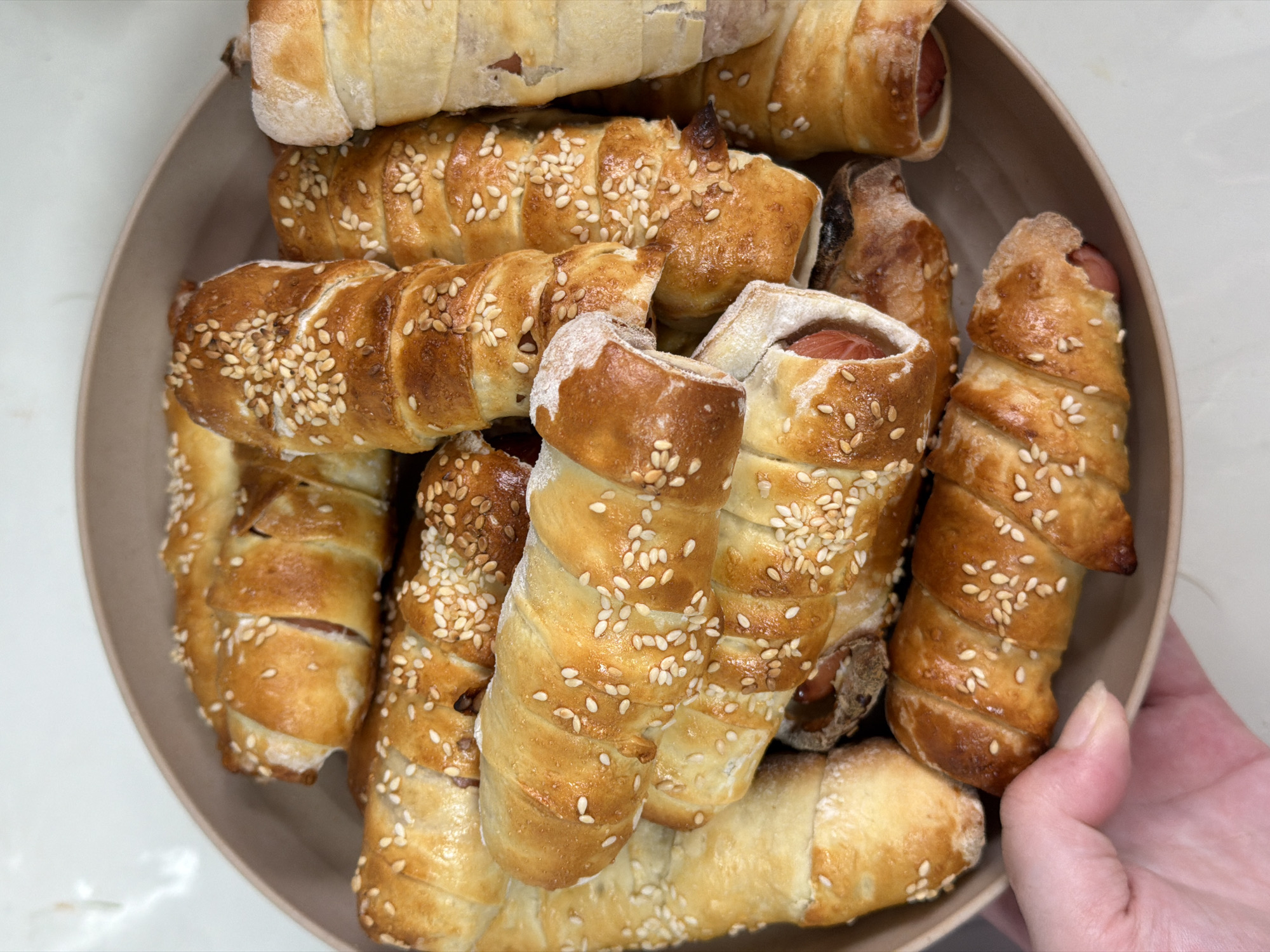 A high-angle, close-up shot shows a bowl filled with several sausage rolls, topped with sesame seeds. A hand is holding the bowl. The background is a light, neutral color. There is no information to suggest the location is the city of Chaikovsky, Russia.