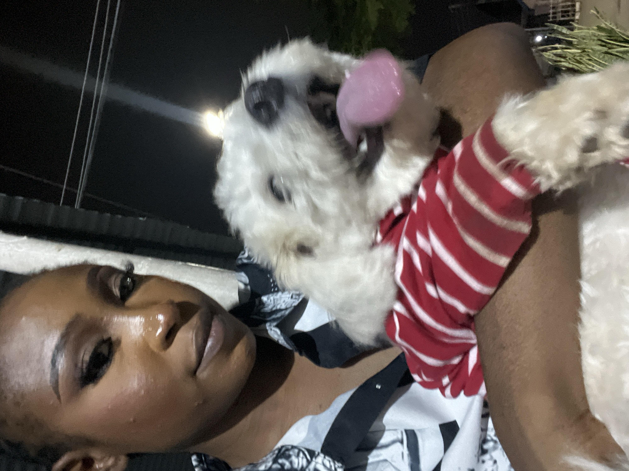 A dark-skinned woman with styled hair and makeup holds a small, white, fluffy dog wearing a red and white striped shirt. The woman is looking at the camera with her mouth slightly open. The dog is looking upwards with its tongue hanging out. They appear to be outdoors at night, with a bright light source in the background. The environment suggests a casual setting in the city of Abuja, Nigeria.