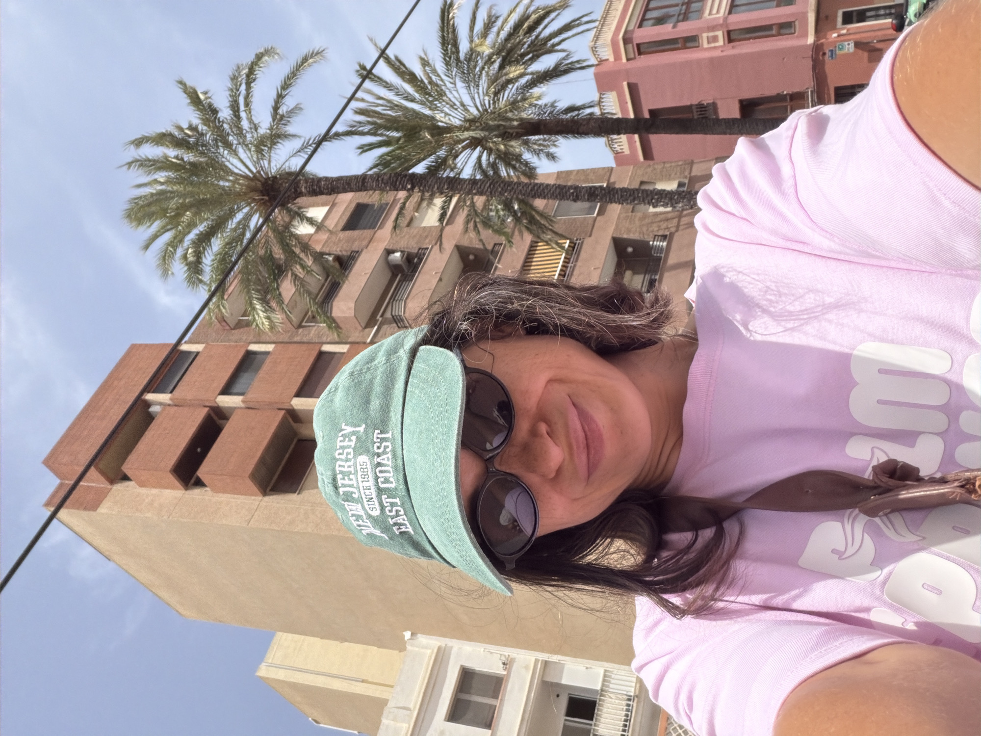 A woman is taking a selfie outdoors in what appears to be a city. She is wearing a green baseball cap that says "NEW JERSEY EAST COAST SINCE 1985", black sunglasses, and a pink t-shirt with white lettering. She has dark hair and is smiling. Behind her are tall buildings and palm trees, suggesting a warm climate. The sky is blue with some scattered clouds.