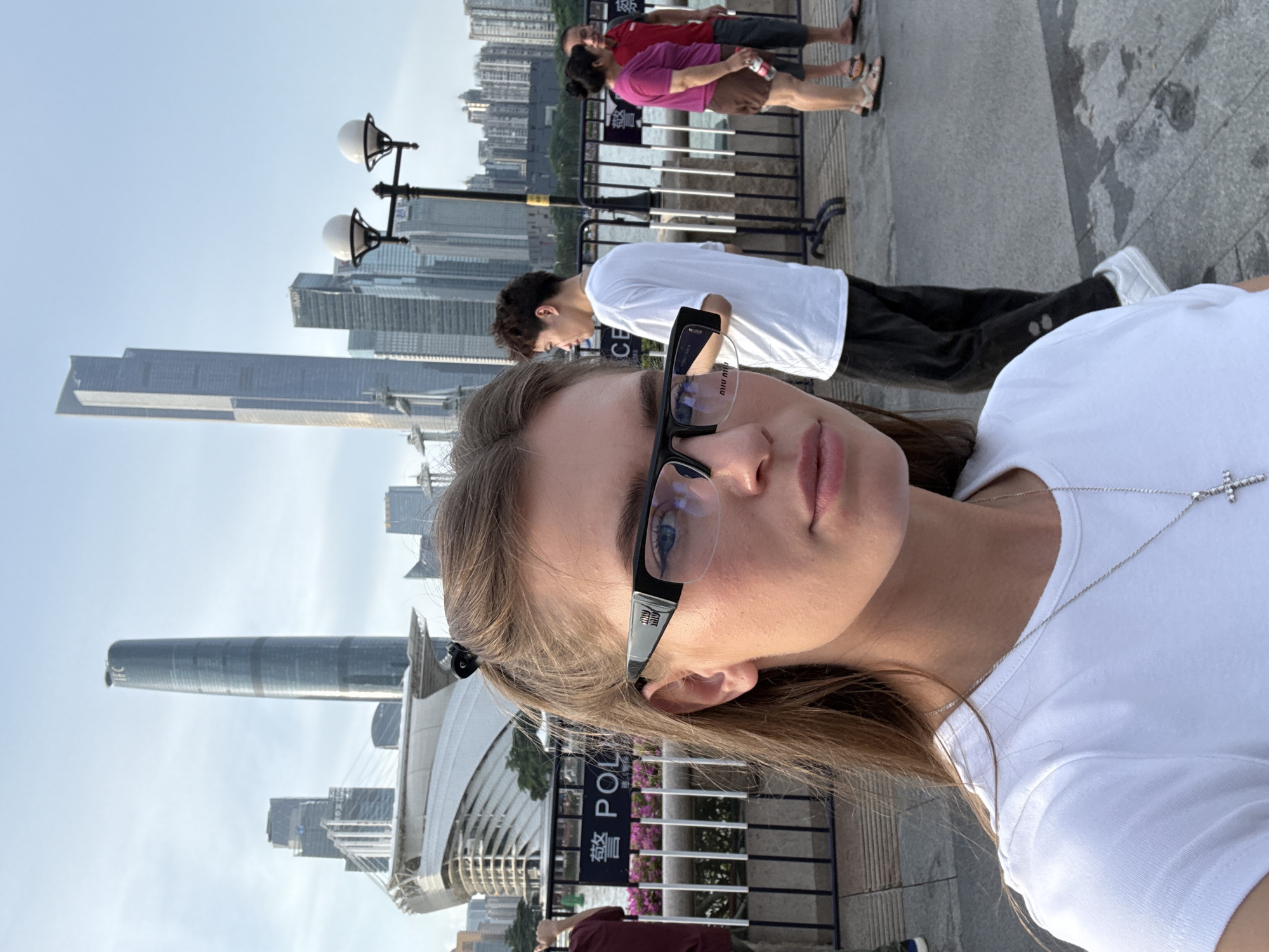 A woman, the primary subject, is captured in a selfie with a shallow depth of field, focusing on her face and upper torso. She is wearing a white t-shirt and a silver necklace with a cross pendant. Her hair is long and brown, and she has on black-rimmed glasses. Behind her, the background is a cityscape with modern architecture, including tall buildings and a distinct cylindrical skyscraper, likely located in Haizhu District, China. The sky is bright and appears clear, suggesting it is daytime with good weather.
In the mid-ground, several other individuals are visible, including a man in a white t-shirt and black pants, and further back, a couple in casual attire. They appear to be walking along a paved walkway or promenade. There are lampposts with globe-shaped lights and a fence line separating the walkway from the buildings.
Visible text includes "POLICE" in both English and Chinese characters on a sign behind the man in the white t-shirt, indicating a potential presence of law enforcement or a regulated area. The overall scene depicts a leisurely day in an urban environment with a scenic waterfront view, implied by the presence of the buildings and promenade. The selfie angle suggests the woman is actively taking the photo of herself.