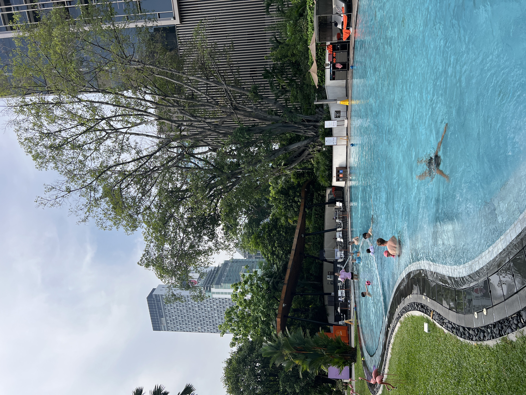 The image shows an outdoor swimming pool area in Bangkok, Thailand. The pool has a curved edge with black decorative stones and a grassy area. Several people are in the pool, including a person swimming laps in the deeper section and a group of people playing near the edge. Surrounding the pool are trees and a building with horizontal stripes. In the background, there is a tall building typical of a city skyline. There is also a table and chairs area next to the pool.