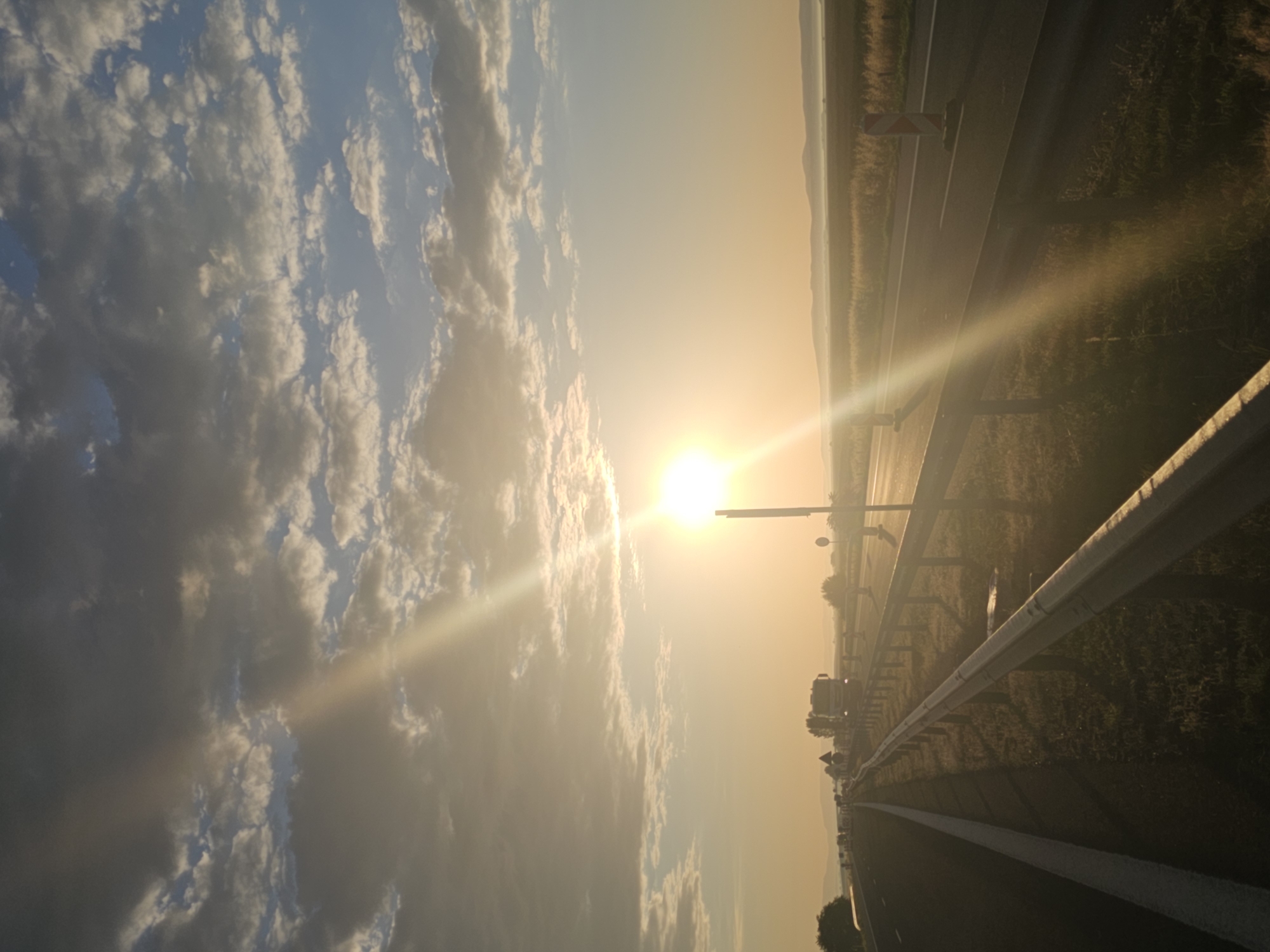 An outdoor, eye-level, vertical, wide shot captures a sunlit highway at dusk. The sun, partially obscured by clouds, beams brightly from the upper center of the frame. The sky is a gradient of warm yellow and orange near the horizon, transitioning to a pale blue at the top, dotted with scattered, textured clouds. A prominent sunbeam cuts diagonally across the scene from the upper left.
The highway itself stretches into the distance, with two lanes visible on the right side of the frame and one lane on the left, separated by a white line. A silver guardrail runs along the right edge of the highway, separating it from a grassy shoulder. In the distance on the highway, several trucks and cars are visible, suggesting traffic. On the right shoulder of the highway, two reflective traffic cones with orange and white stripes stand out. The landscape beyond the highway appears flat, with fields of dry grass and distant, hazy hills. The lighting suggests either sunrise or sunset, casting long shadows. The image appears to be taken in or around the village of Zagortsi, Bulgaria, given the context of the provided location.