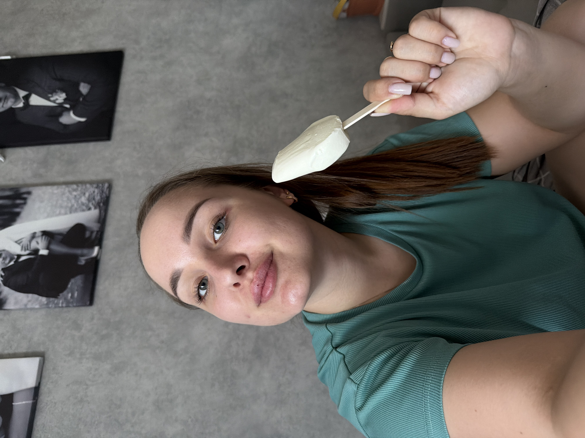 A woman is holding a white ice cream bar on a wooden stick in her right hand. She is looking at the camera. She is wearing a green shirt. Behind her are black and white photos mounted on a gray wall. It is not possible to determine the location of Chaikovsky, Russia.