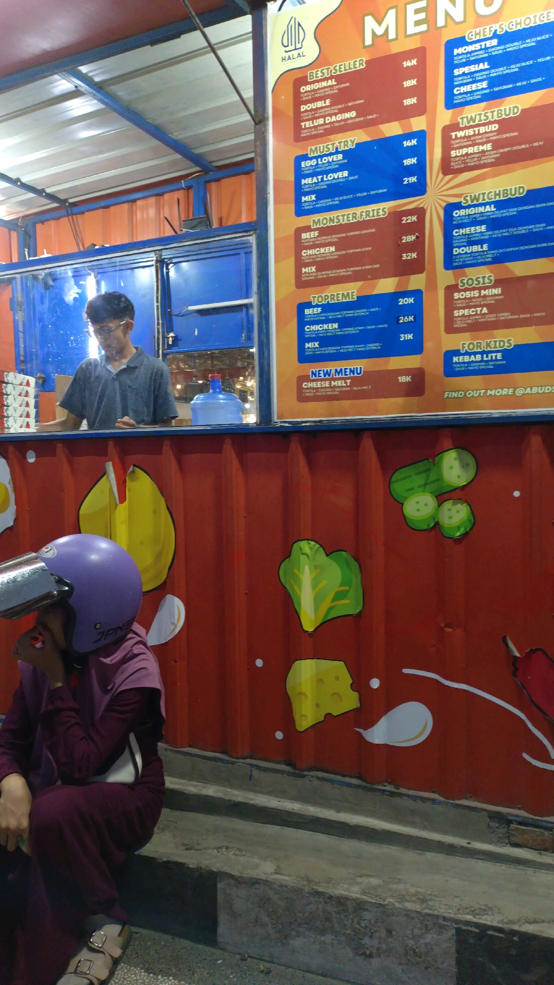 In Padang Panjang, Indonesia, a person wearing a purple hijab and a maroon outfit sits on a concrete step in front of a red corrugated metal structure. They have a purple helmet resting on their head and are looking down. Above them, a menu board displays various food items and prices. A man in glasses is visible behind a blue serving counter, appearing to interact with customers. The wall behind the person is decorated with cartoonish drawings of food items like cheese, lettuce, and cucumbers. The overall scene depicts a street food vendor setting.