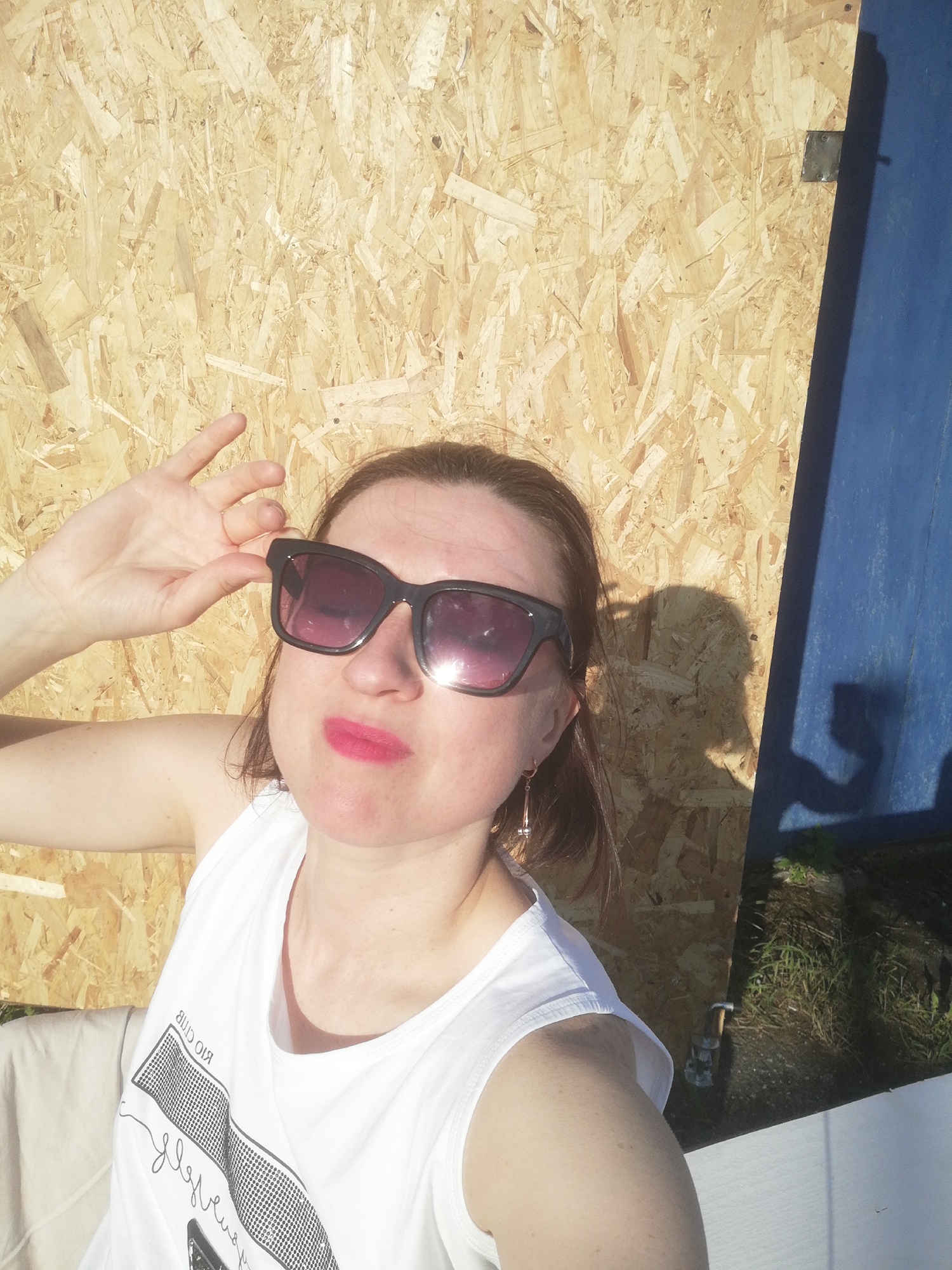 An overhead medium shot captures a woman in a white tank top and sunglasses, her left hand adjusting the sunglasses. She has bright pink lipstick. The background is a textured wooden wall. To the right of the woman, a dark blue vertical surface is visible, casting shadows. The scene appears to be outdoors, with the suggestion of grass and dirt on the ground in the lower right corner. The image does not provide specific landmarks or indications of the city of Tosno, Russia.