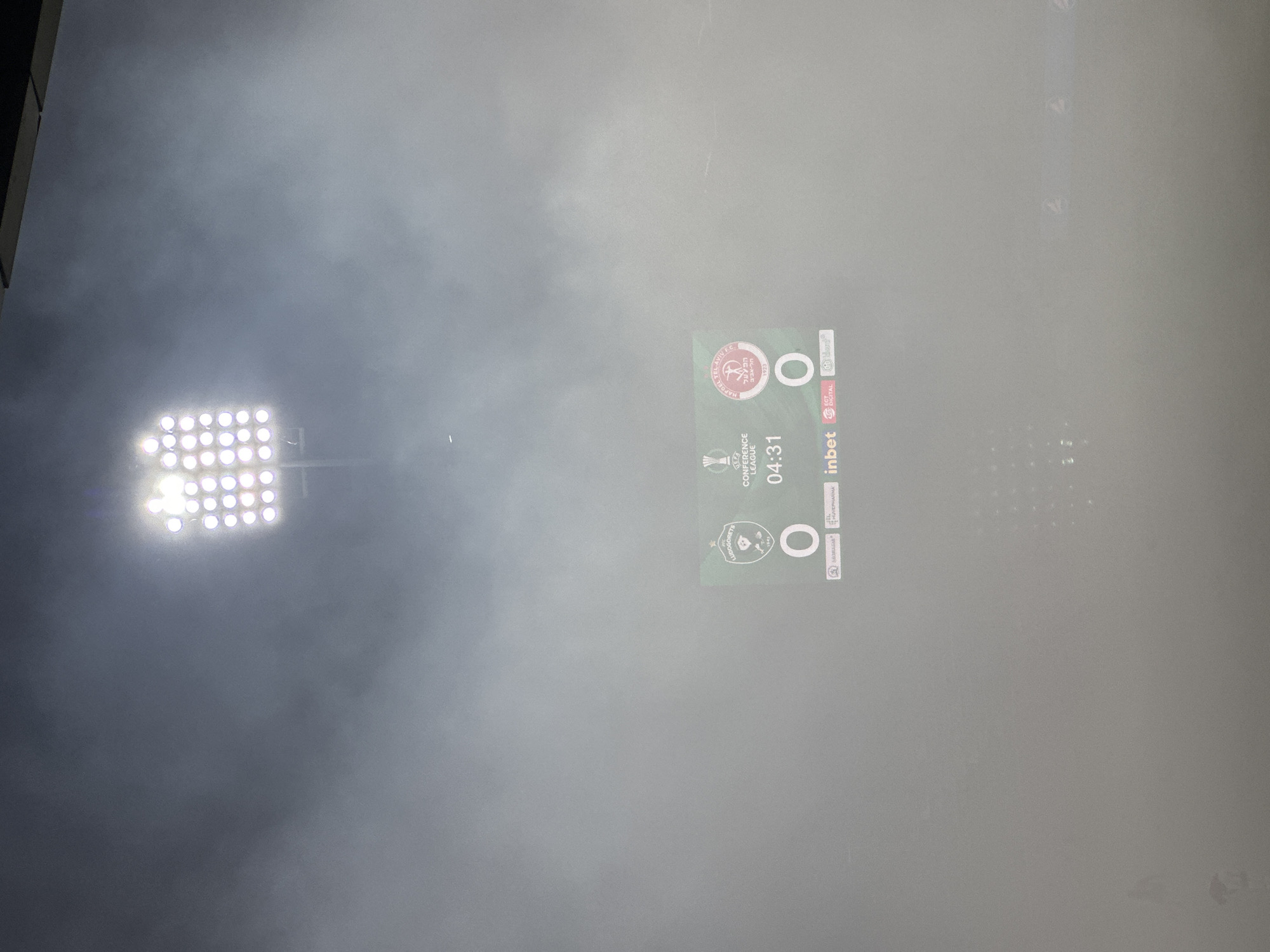 A foggy outdoor stadium environment is depicted, with a large floodlight illuminating the scene from above. A digital scoreboard is visible in the center, displaying a football match score. The scoreboard shows "0" for both teams and a timer reading "04:31". The UEFA Europa Conference League logo is present on the scoreboard, along with team crests and sponsor logos. The stadium seating area is dimly visible in the background, with a faint pattern of lights suggesting a grid-like structure. The overall atmosphere is hazy and somewhat indistinct due to the fog.