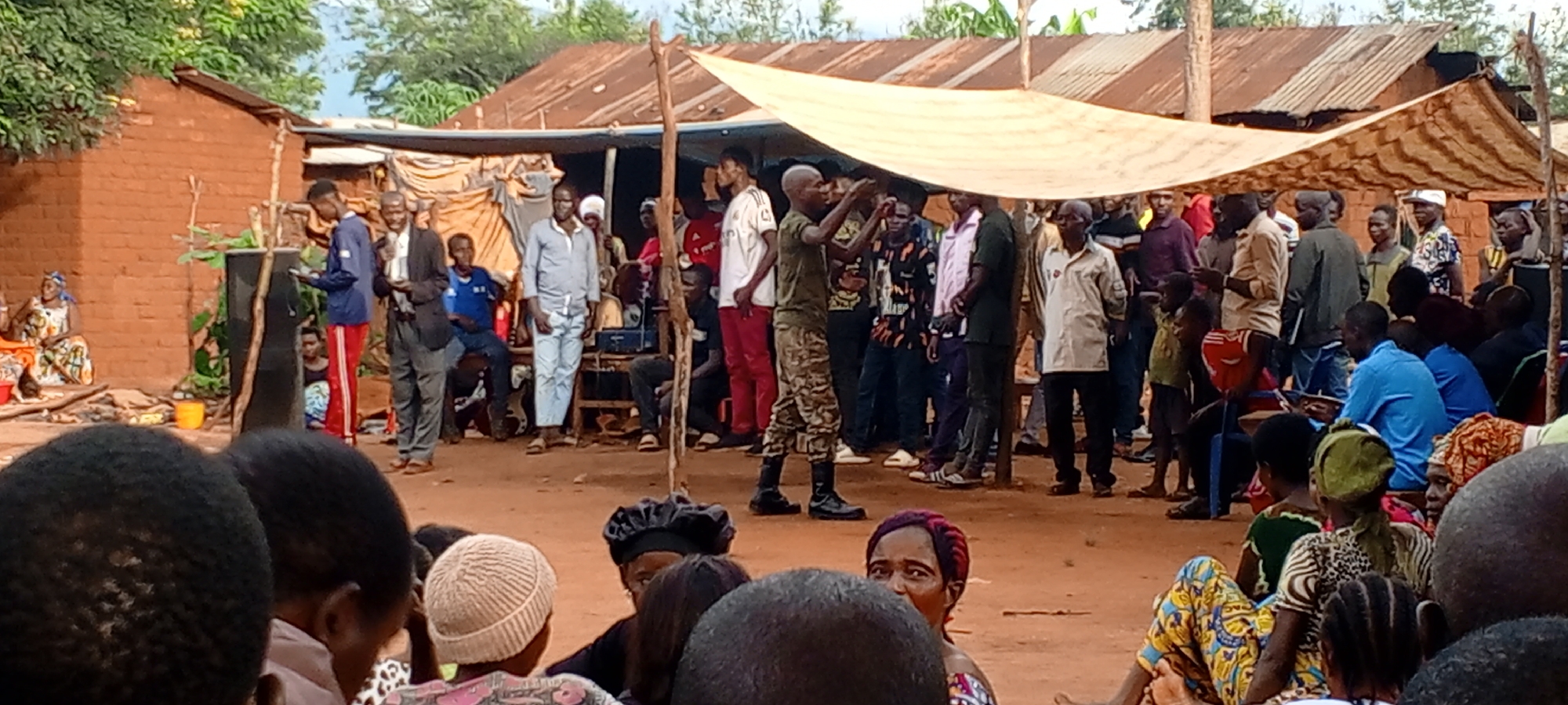 The image depicts an outdoor gathering of people in Ngalula, Democratic Republic of the Congo. A large group of individuals is seen standing and sitting in a dirt courtyard. A makeshift shelter is erected above them. Several people stand near a speaker's podium. A person in military uniform is gesturing with their hands. Some people are seated on benches and the ground in the foreground, observing the scene. There is a brick building behind some of the people.