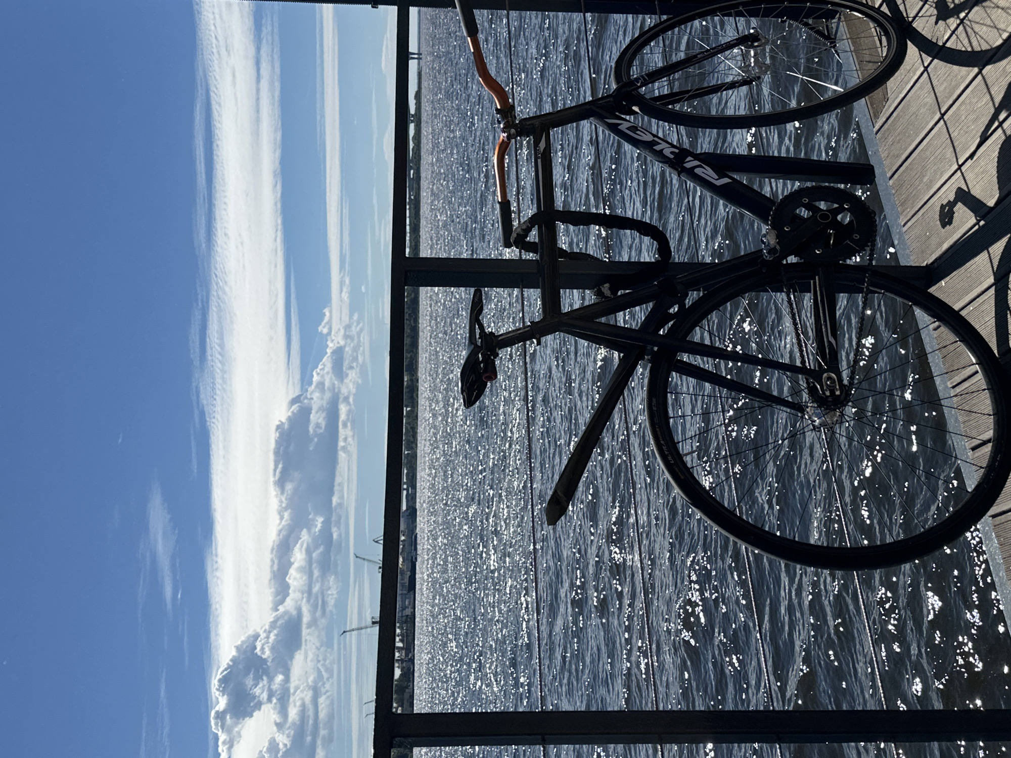 A black bicycle is parked on a wooden pier beside a railing, overlooking a body of shimmering water. The water is a deep blue, reflecting the bright sunlight and the sky above. In the distance, industrial cranes and a bridge are visible along the shoreline, suggesting an urban industrial area. The sky is a vibrant blue with scattered white clouds, some of which are wispy and stretched horizontally, while others are larger and more voluminous. The scene is set in Saint Petersburg, Russia, likely along the Neva River or one of its tributaries, given the industrial landscape and expansive water views characteristic of the city's waterfront. There are no people present in the image. The bicycle is locked to the railing with a thick chain. The sun casts a distinct shadow of the bicycle and railing onto the wooden planks of the pier.