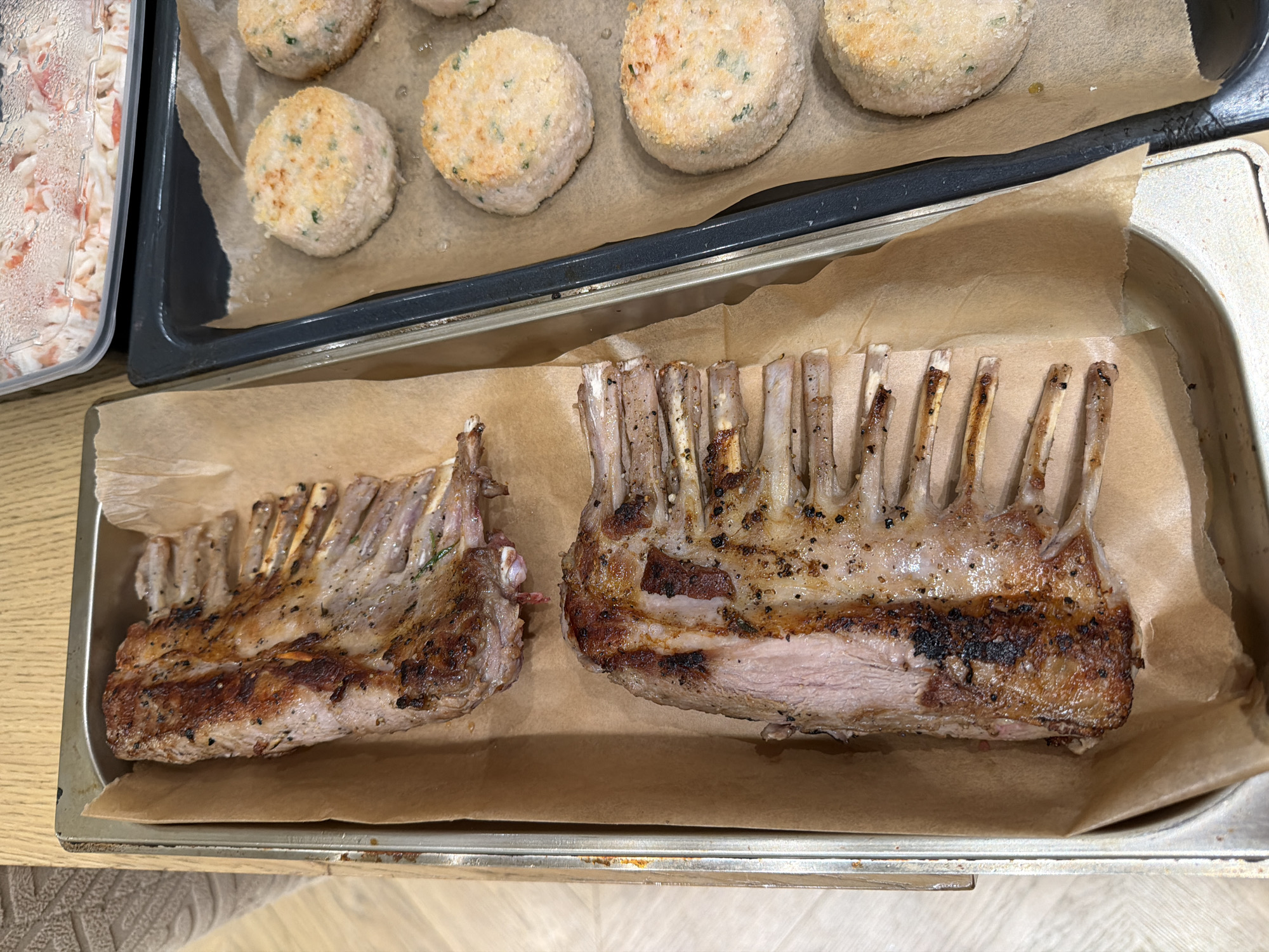 The image depicts a close-up, overhead view of two racks of seasoned and roasted lamb chops arranged on parchment paper within a stainless steel tray. The lamb chops are a rich brown color with visible seasoning, and the meat appears to be cooked to a medium-rare to medium doneness. To the right of the lamb, on a dark baking sheet also lined with parchment paper, are several round, breaded patties, likely fish or meat cakes. The background shows a portion of a wooden surface and a clear plastic container filled with a pink and white food item, possibly a seafood salad. The overall setting suggests a kitchen or preparation area. There are no discernible elements in the image that specifically indicate a location in Moscow, Russia.