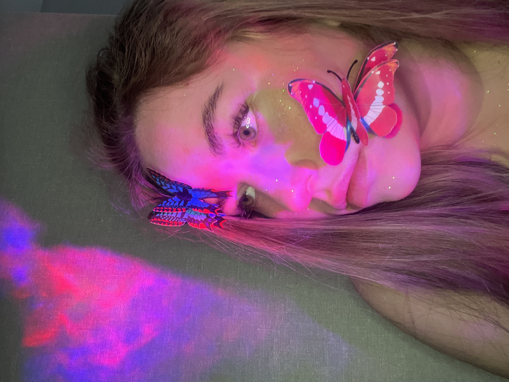 A young woman with light brown hair lies on a gray surface. Two artificial butterflies are attached to her face: a red one near her cheek and a blue one near her forehead. There appears to be glitter on her face. The scene is bathed in purple and pink light, and the location cannot be determined from the image.